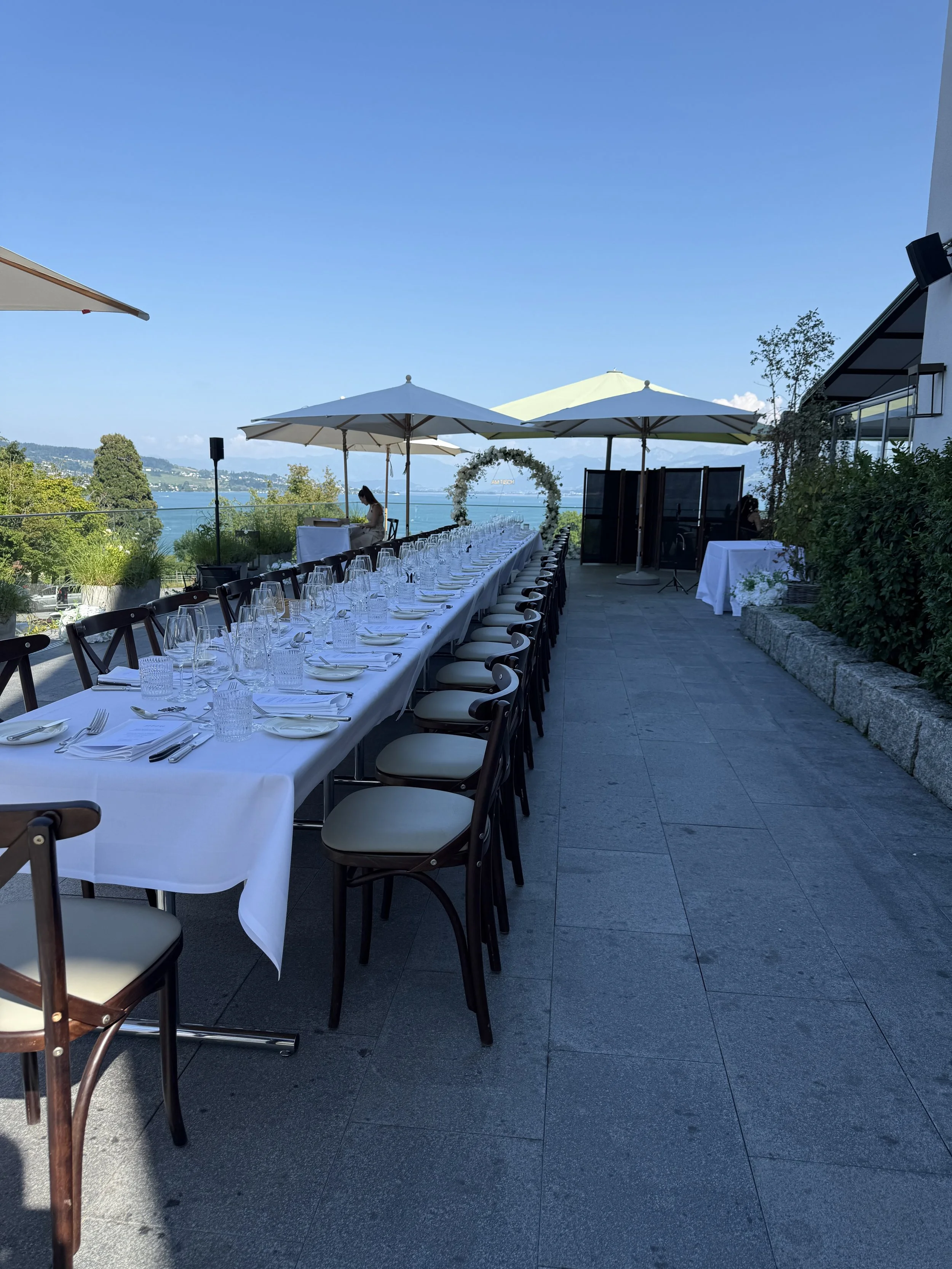 Ein festlich gedeckter Tisch mit weißen Tuch, Wasser- und Weingläsern sowie Besteck, auf einer Terrasse mit Blick auf den See, im Hintergrund sind Bäume und Berge sichtbar, unter Sonnenschirmen im Freien