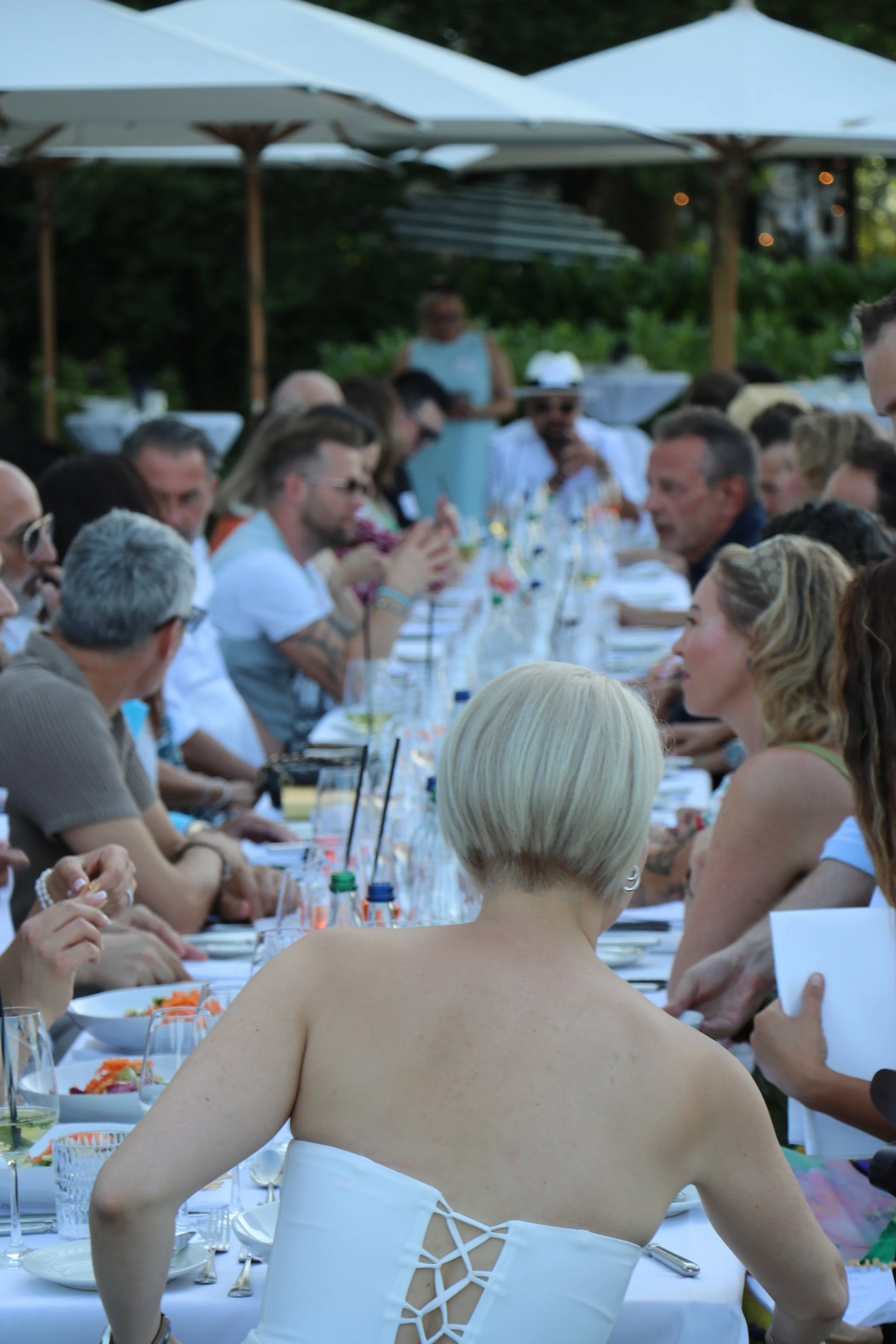 Menschen sitzen an einem langen Tisch im Freien bei einem Dinner, unter Sonnenschirmen, in einer grünen Gartenumgebung.