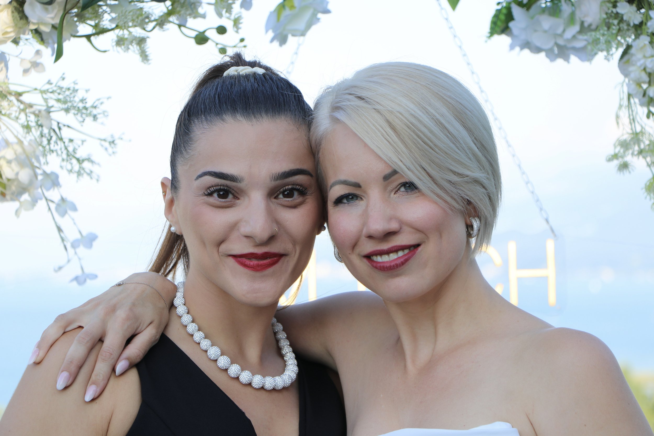 Zwei Frauen umarmen sich lächelnd bei einer Hochzeit im Freien, blauer Himmel, Blumen im Hintergrund.