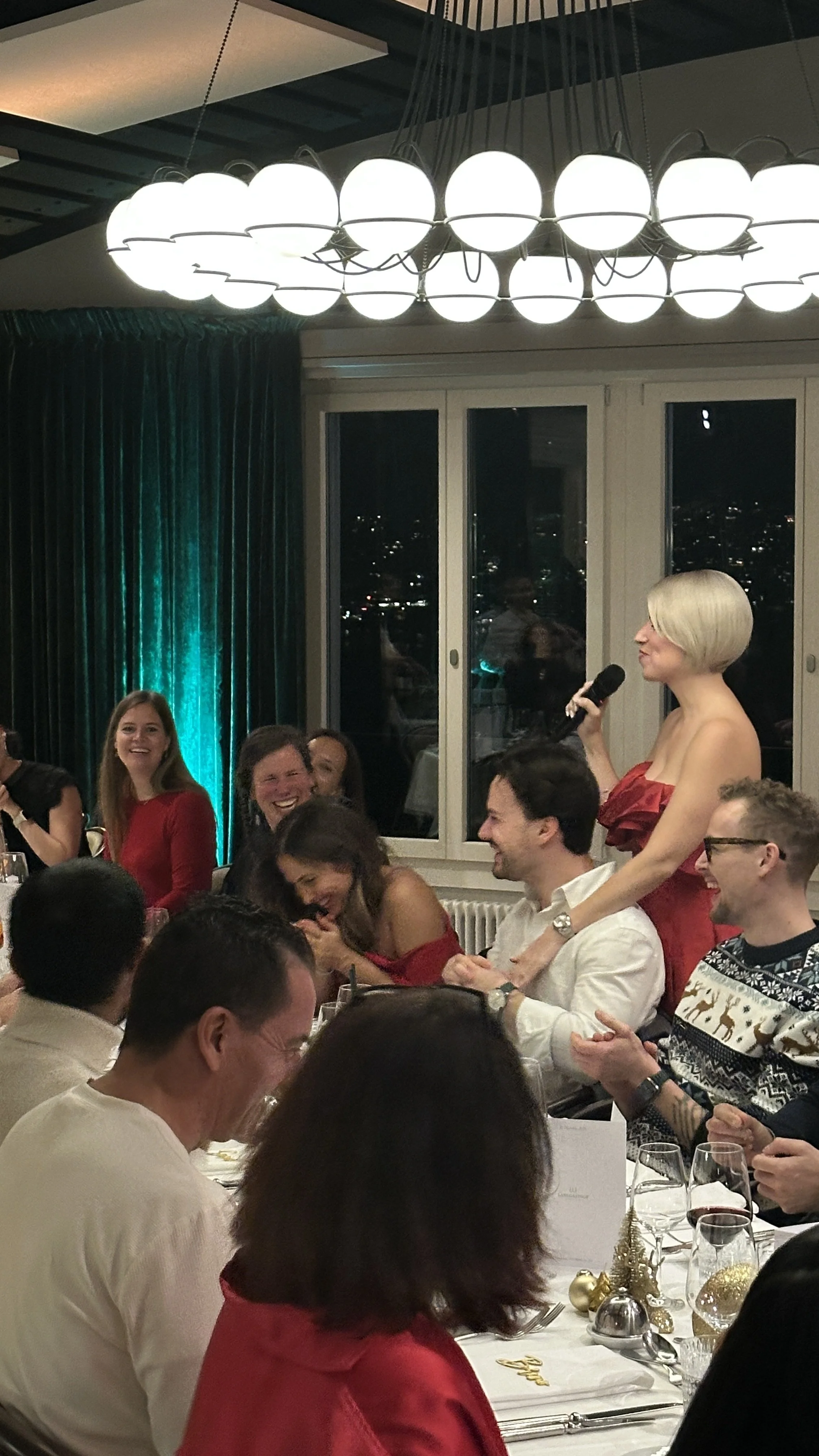 Menschen feiern bei einem Festessen, eine Frau mit kurzem blondem Haar hält Mikrofon und spricht, die Gäste lachen und haben Spaß.