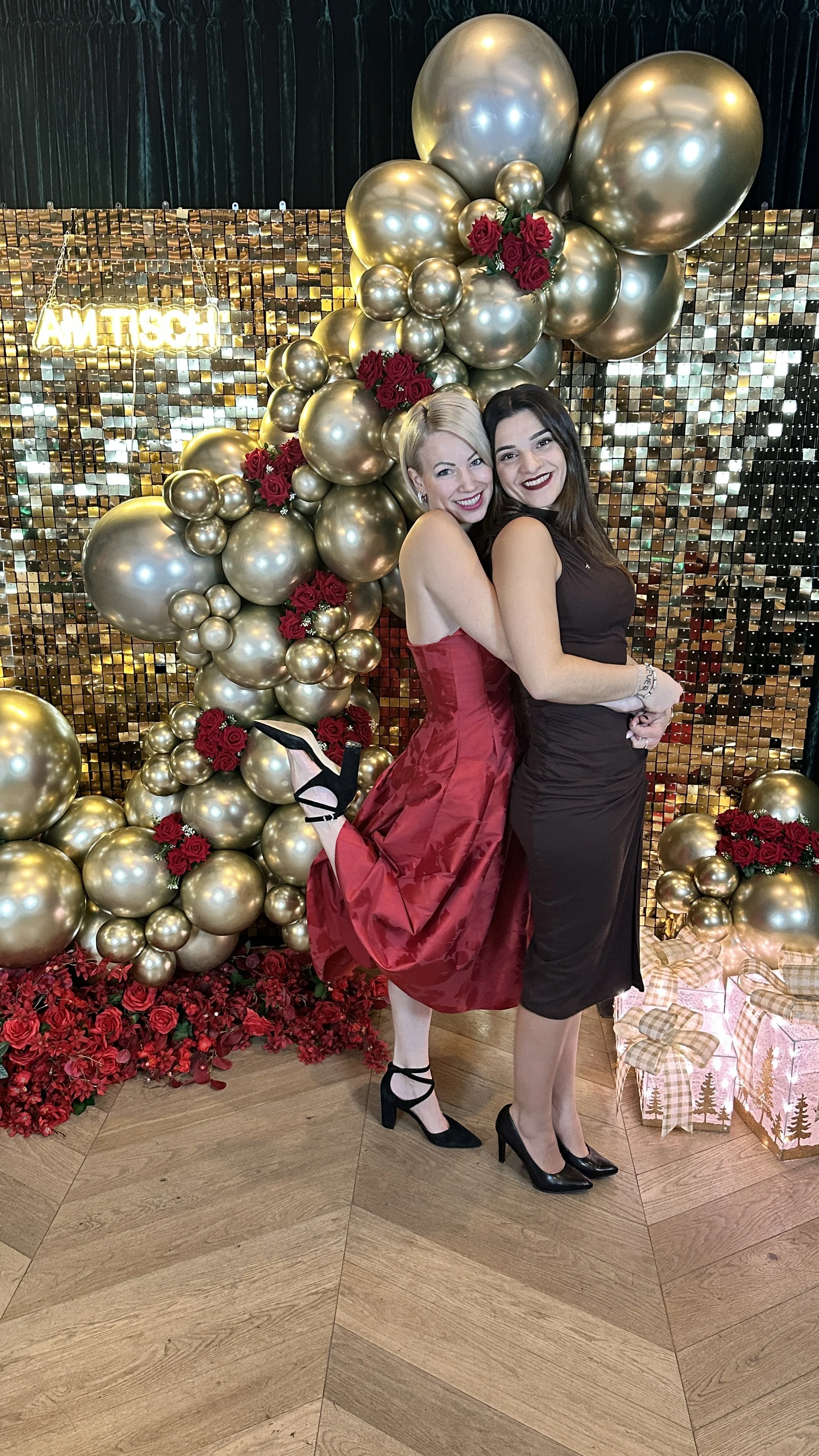 Zwei lachende Frauen in eleganten Kleidern vor einer gold-braun Dekoration mit Ballons und Blumen bei einer Feier.