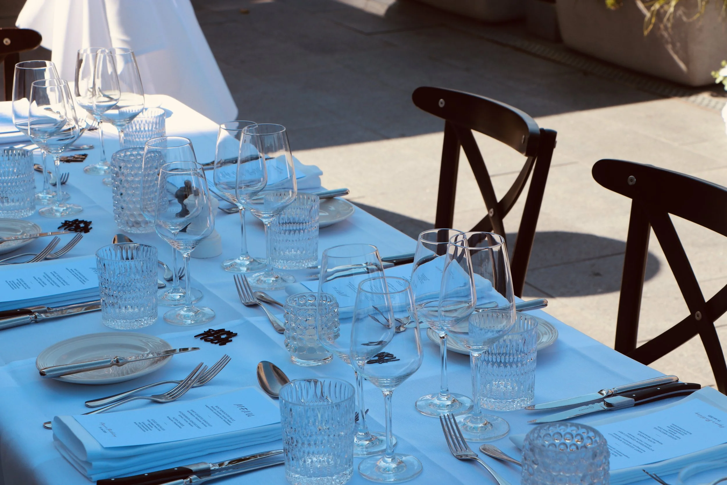 Elegant gedeckter Tisch für ein Essen mit weißen Servietten, zahlreichen Weingläsern, Wasser Gläsern, Tellern, Besteck und Menükarten. Der Tisch steht im Freien, mit Schatten und Sonnenschein auf einer Terrasse.