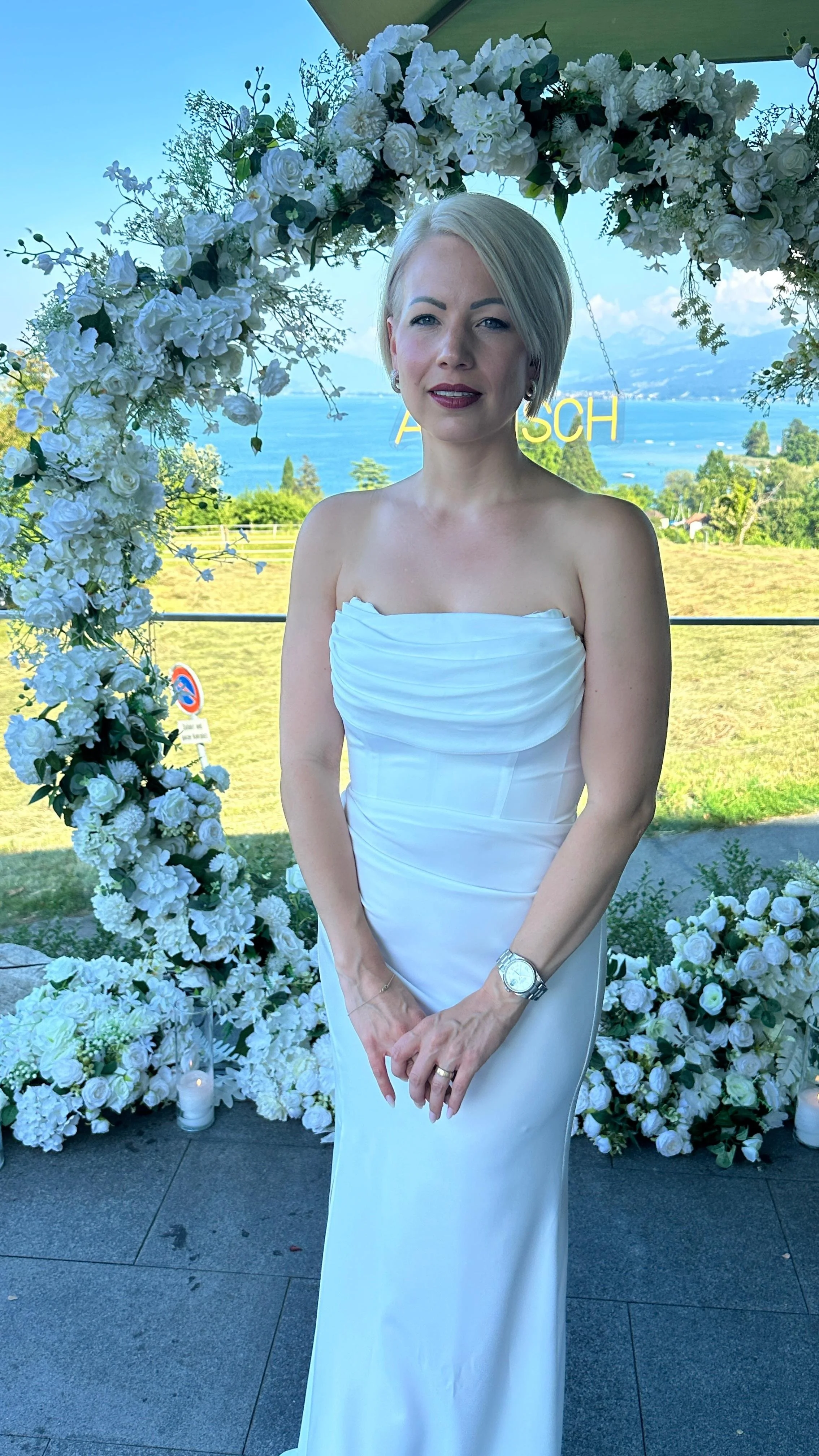 Frau in weißem Hochzeitskleid vor einer blumenreichen Kulisse und einer Wasserlandschaft im Hintergrund bei einer Hochzeit.