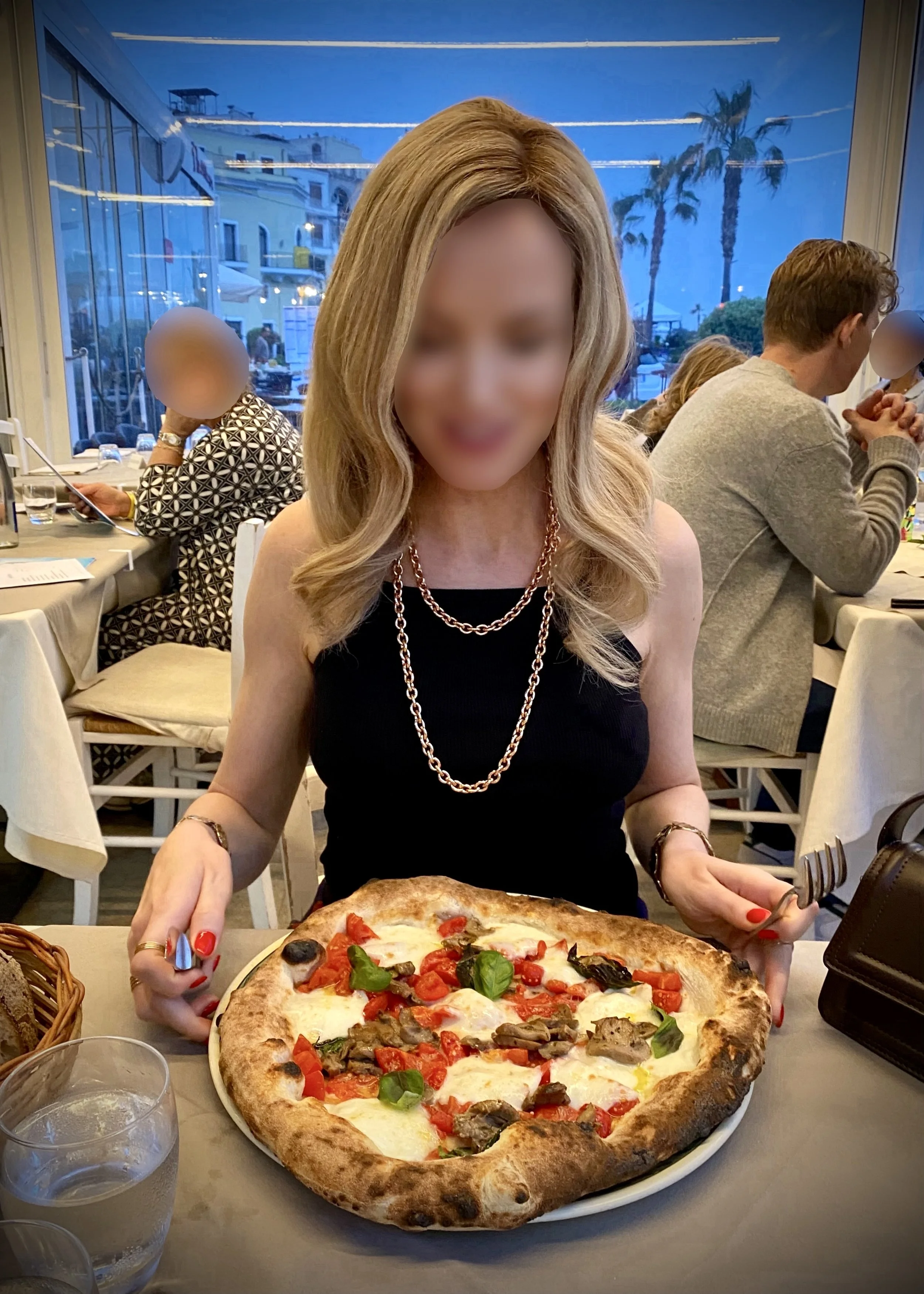Blonde high class independent London escort sitting at a restaurant table with a large pizza topped with cheese, cherry tomatoes, basil, and mushrooms, holding a fork and knife. Other diners are visible in the background.