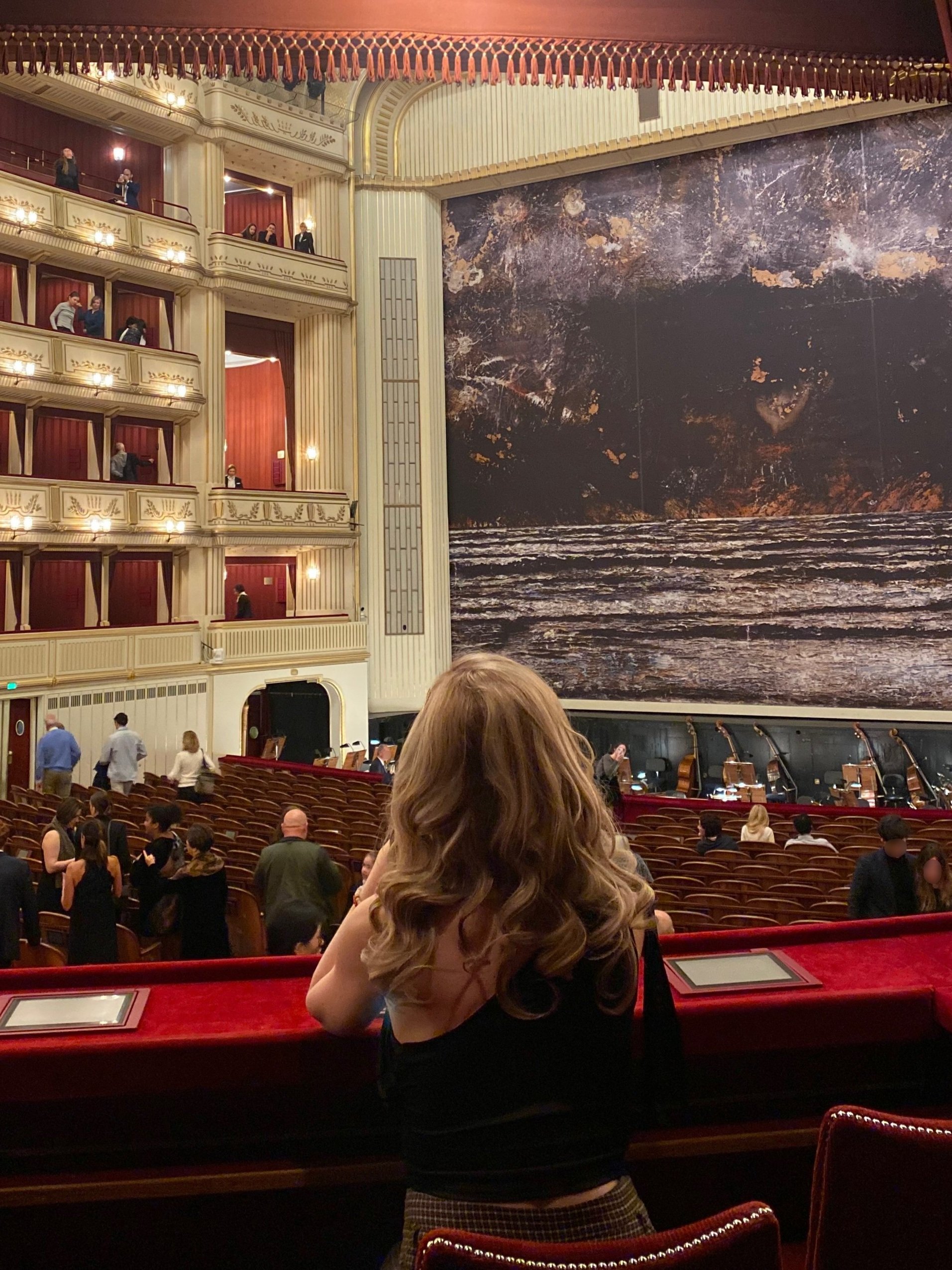 Interior of the Vienna Opera House with balconies and red velvet seating, featuring a stage with a large, textured backdrop. A blonde high class independent London escort is observing the scene from a box.
