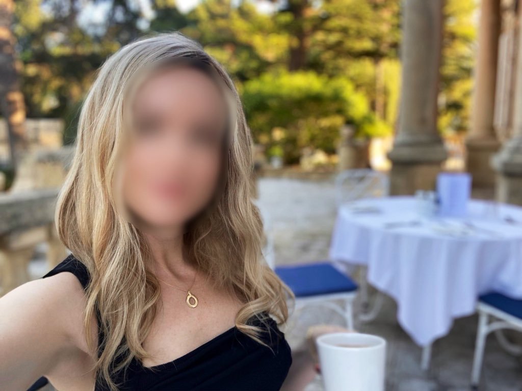 Outdoor scene with a blonde high class independent London escort wearing a black sleeveless top, seated at a table set for dining.