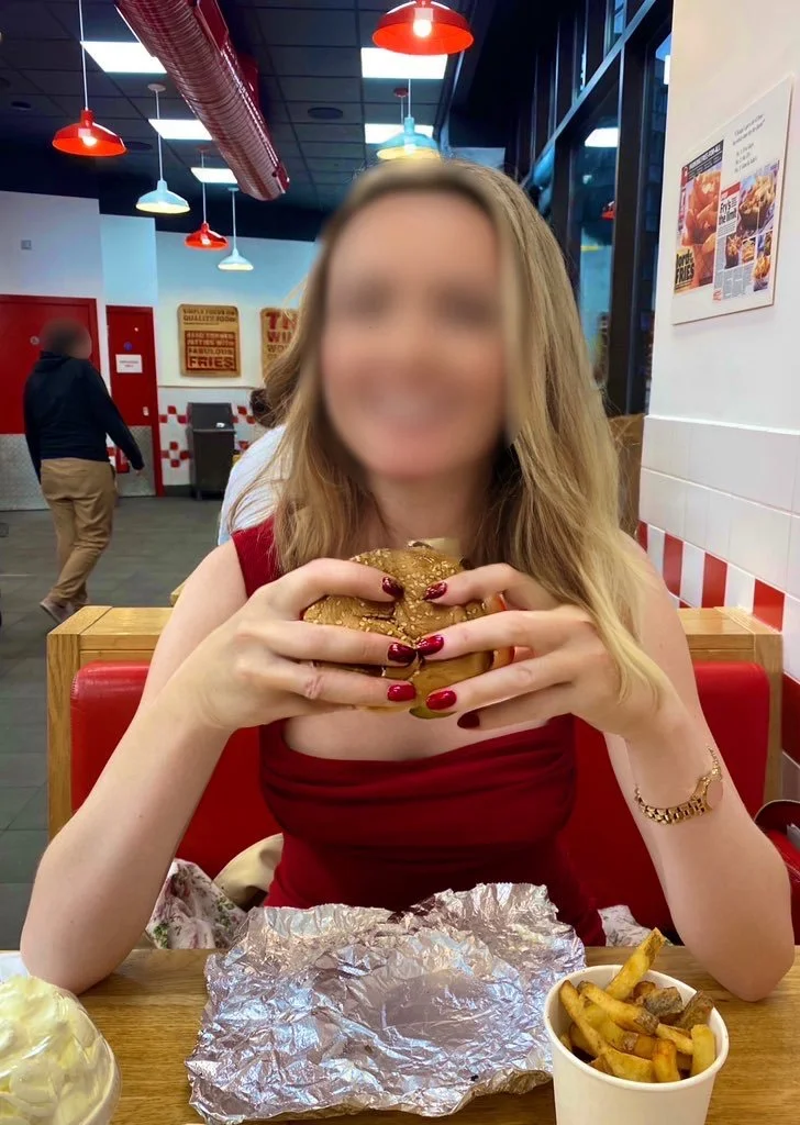 A blonde high class independent London escort in a Five Guys fast-food restaurant holding a burger, on a lunch date.