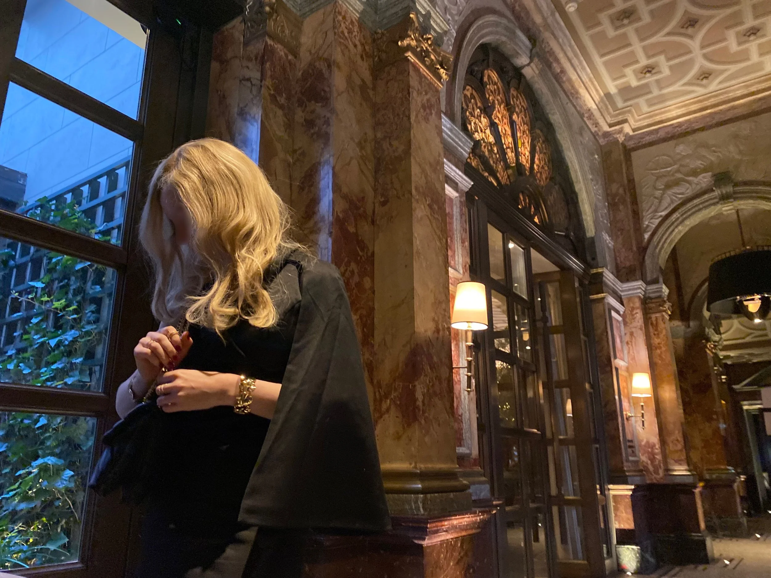 Blonde high class independent London escort by a window, in a luxurious setting with ornate marble and wood paneling, wearing an elegant black evening dress.