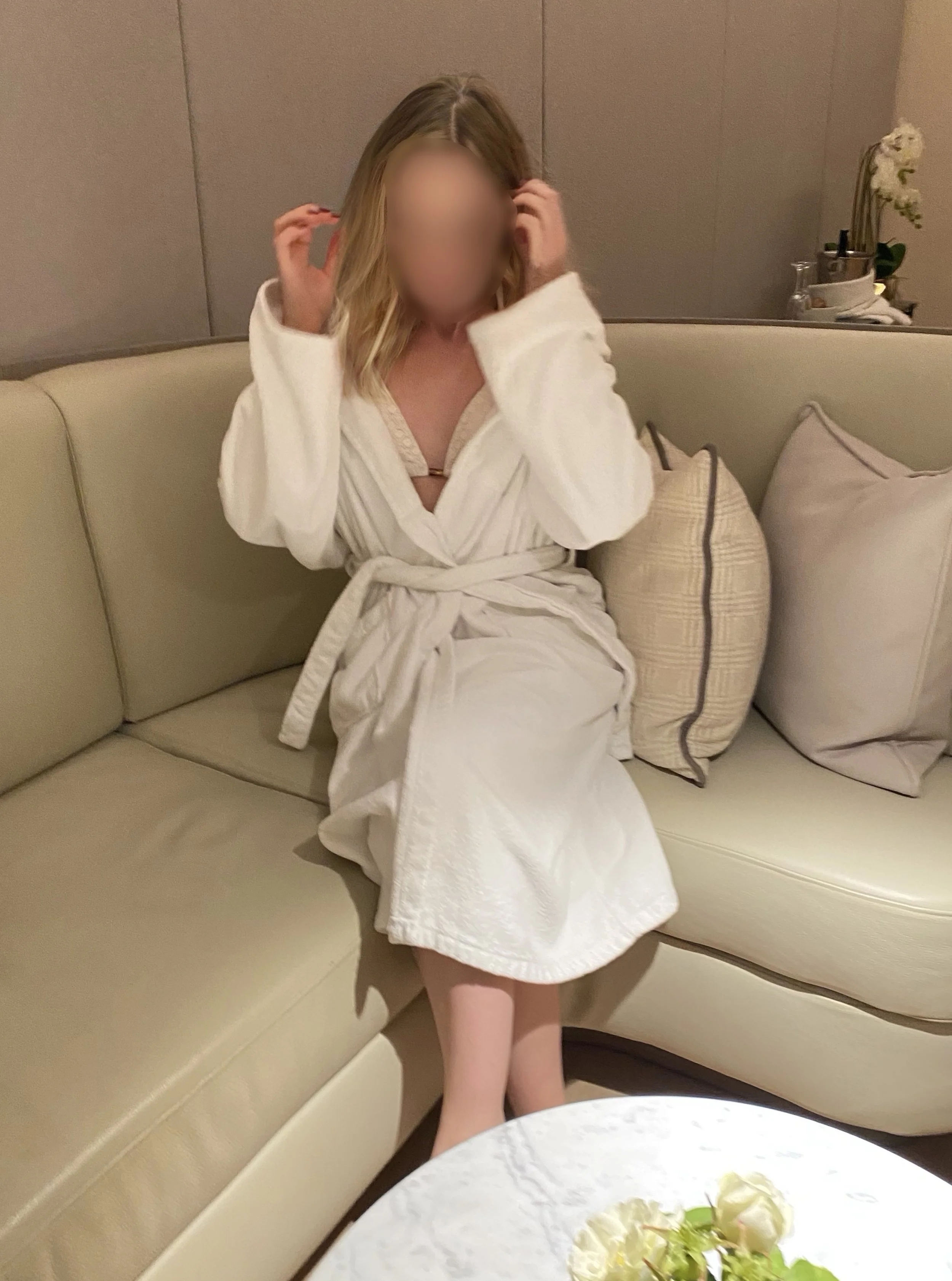 Blonde high class independent London escort sitting on a couch in a luxury spa wearing a white bathrobe, with blurred face, adjusting her hair.