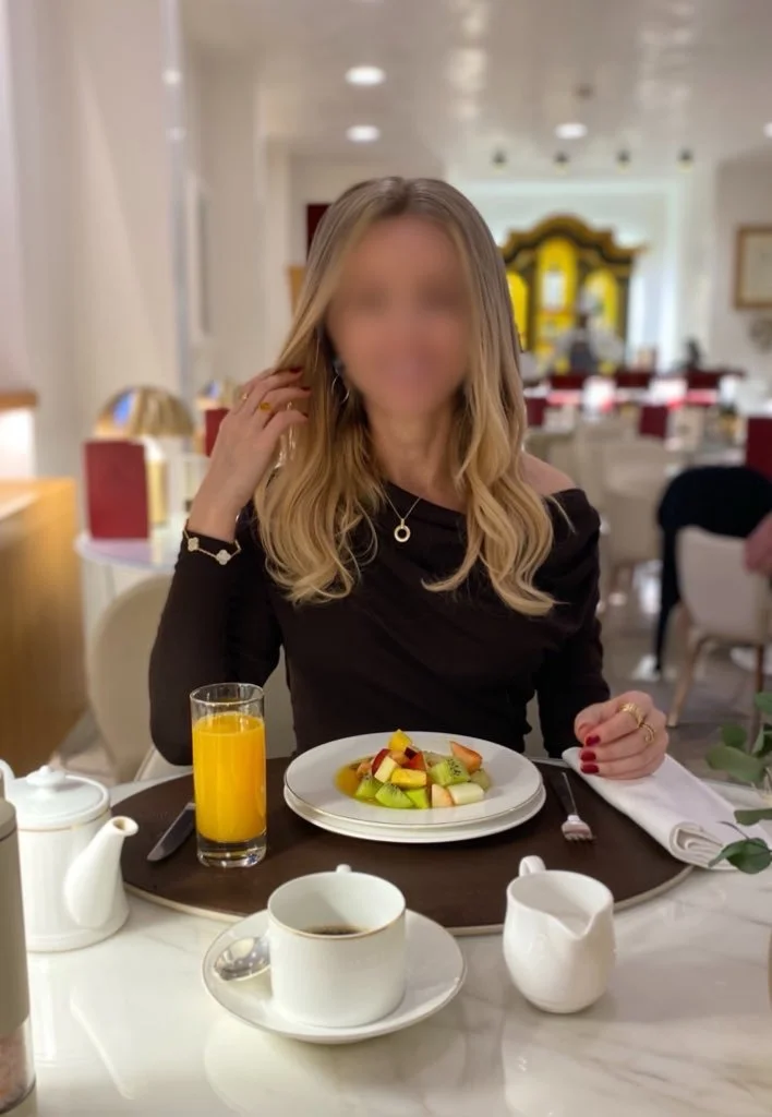 Blonde high class independent London escort sitting at a dining table with fruit salad and orange juice, after an overnight FMTY date.