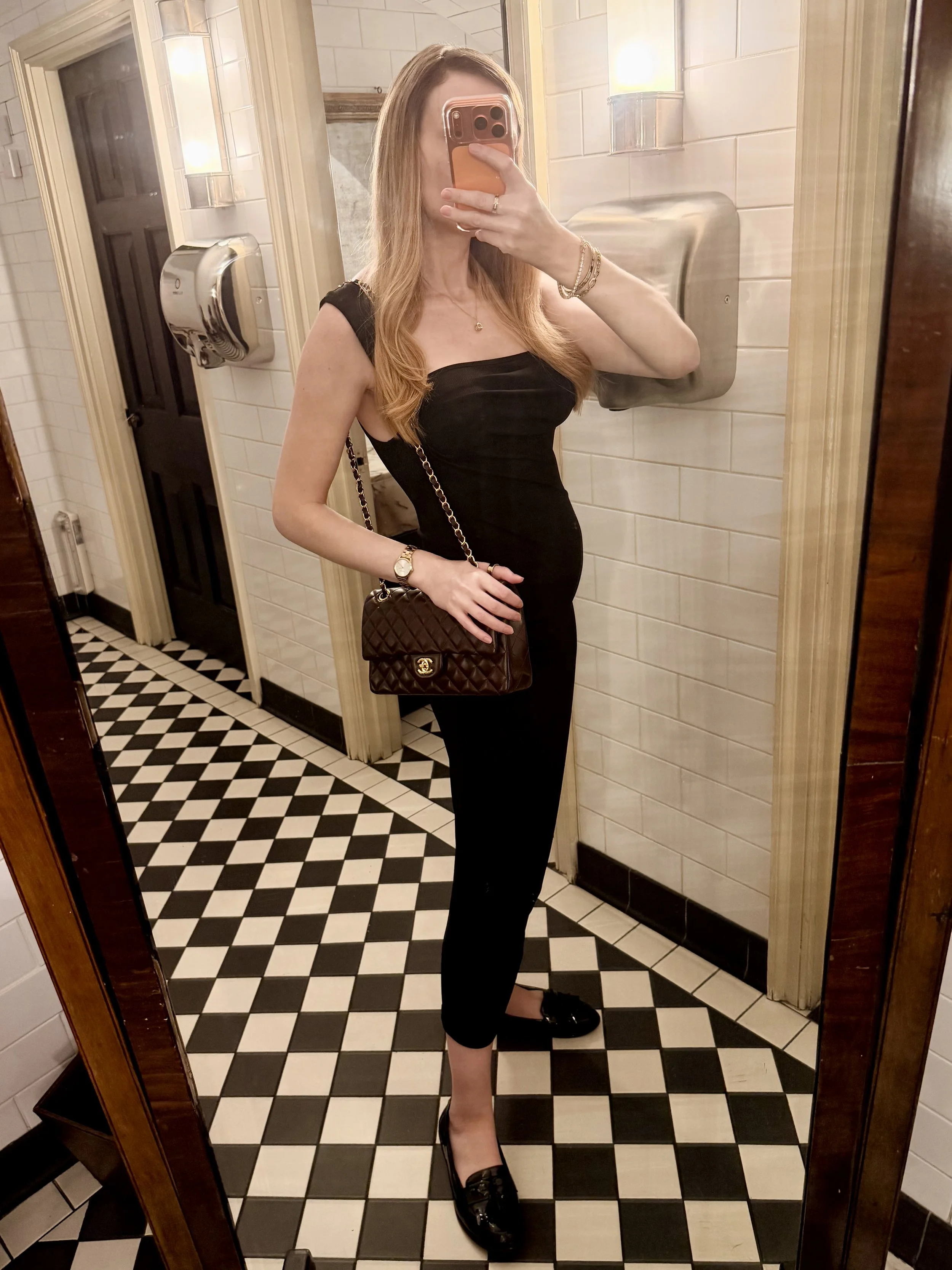 An independent pregnant luxury escort is taking a mirror selfie in a London bathroom, wearing a black sleeveless dress.