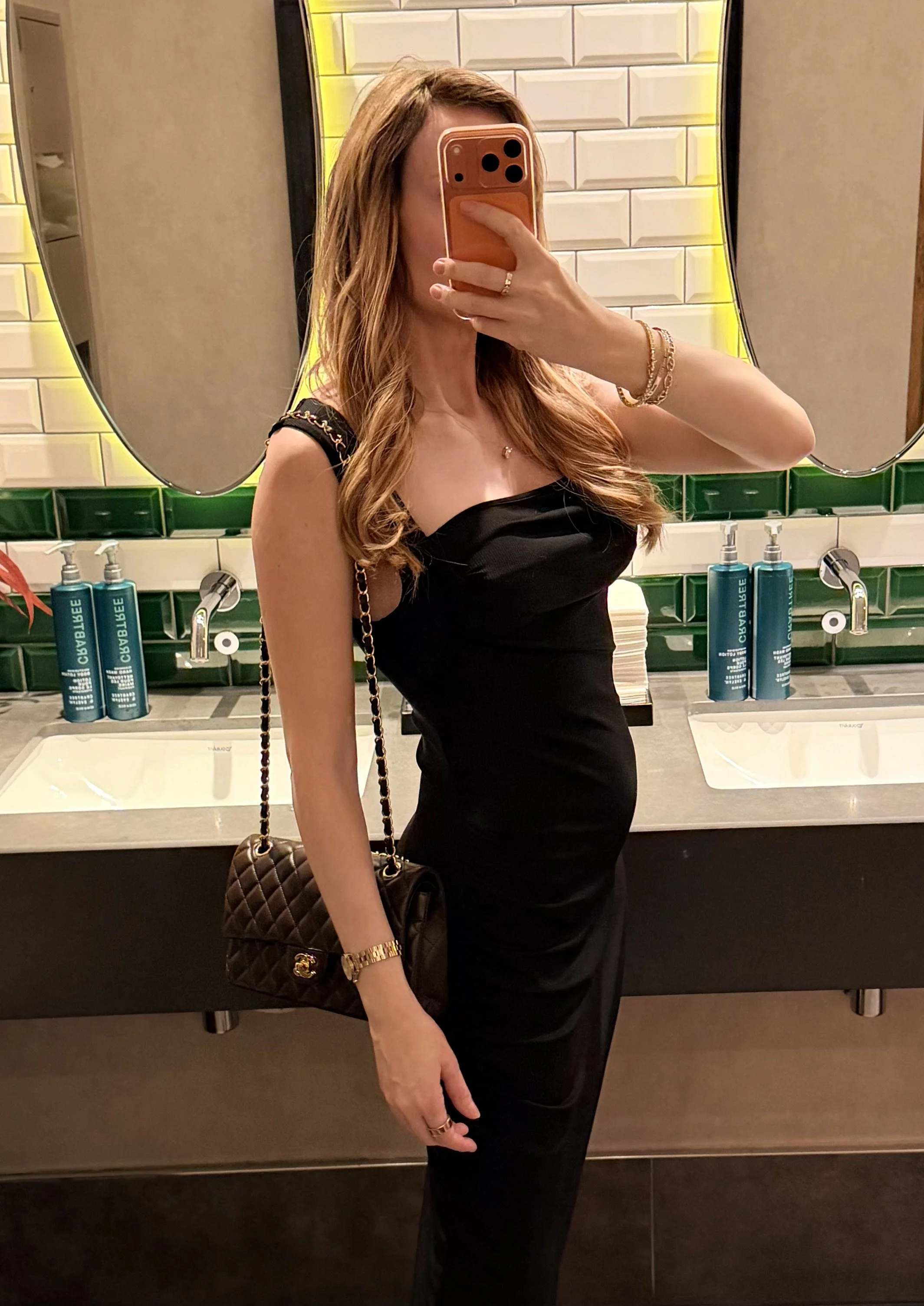 A pregnant GFE escort taking a mirror selfie in a bathroom, dressed in a black dress, carrying a quilted Chanel purse with a chain strap, wearing jewelry, with sinks and soap bottles in the background.