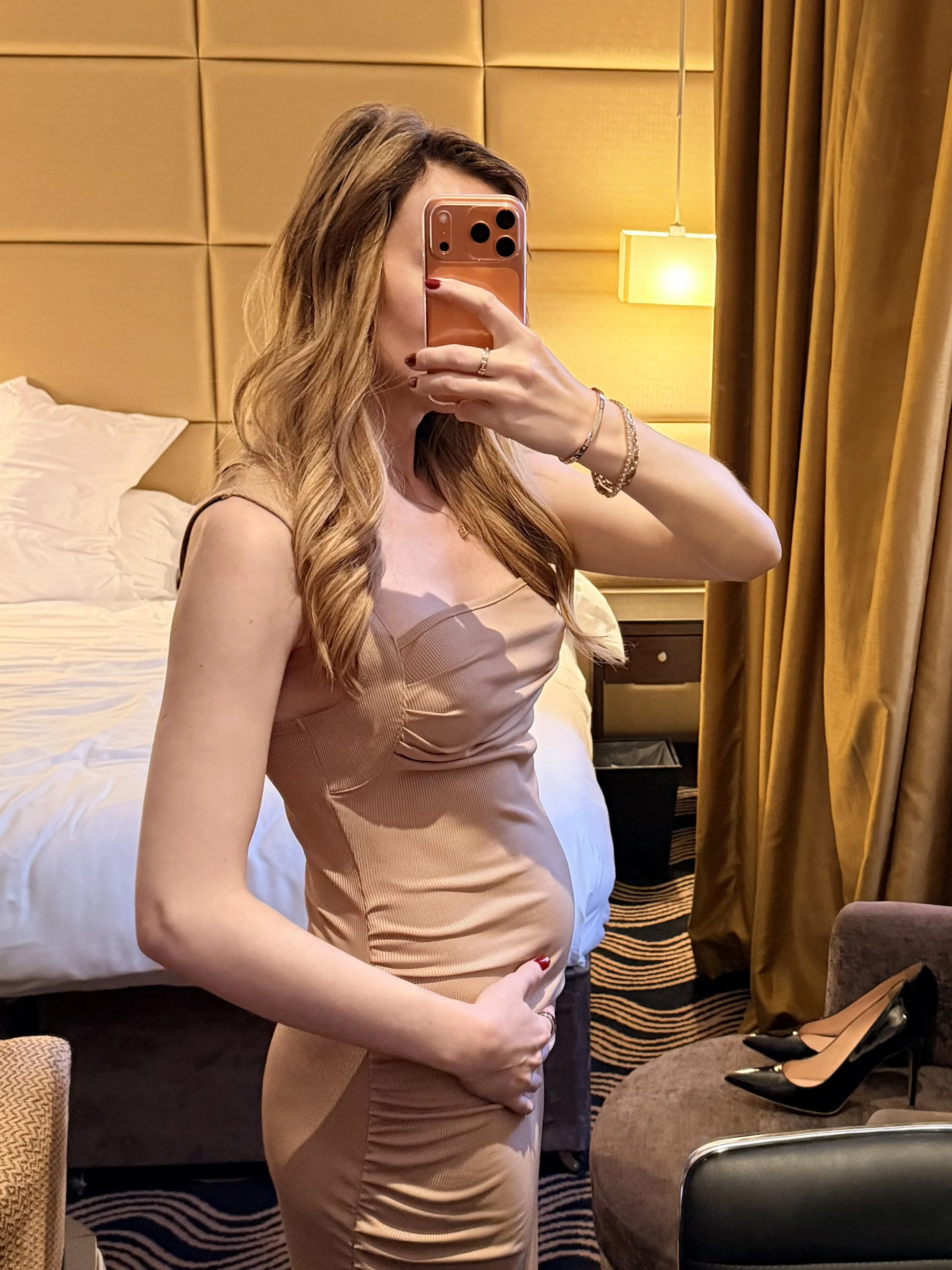 A London independent pregnant companion taking a mirror selfie in a hotel room, holding her round belly with one hand, wearing a beige dress, with shoes on a chair nearby.