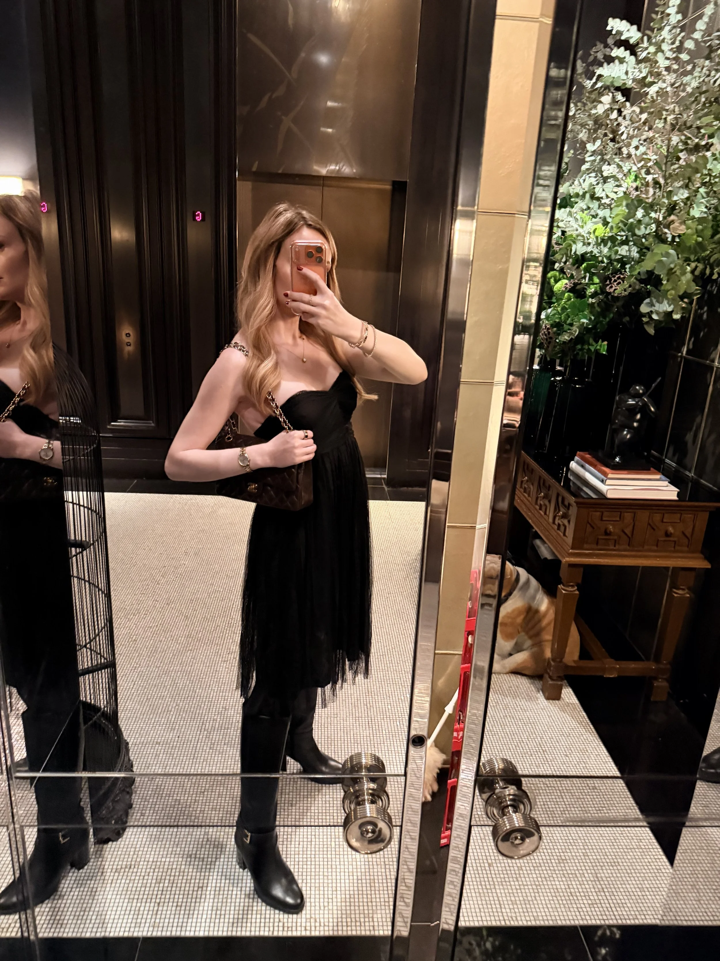 A pregnant high class escort taking a mirror selfie in an elevator, wearing a black dress with a tulle skirt, black knee-high boots, and holding a brown Chanel handbag. 