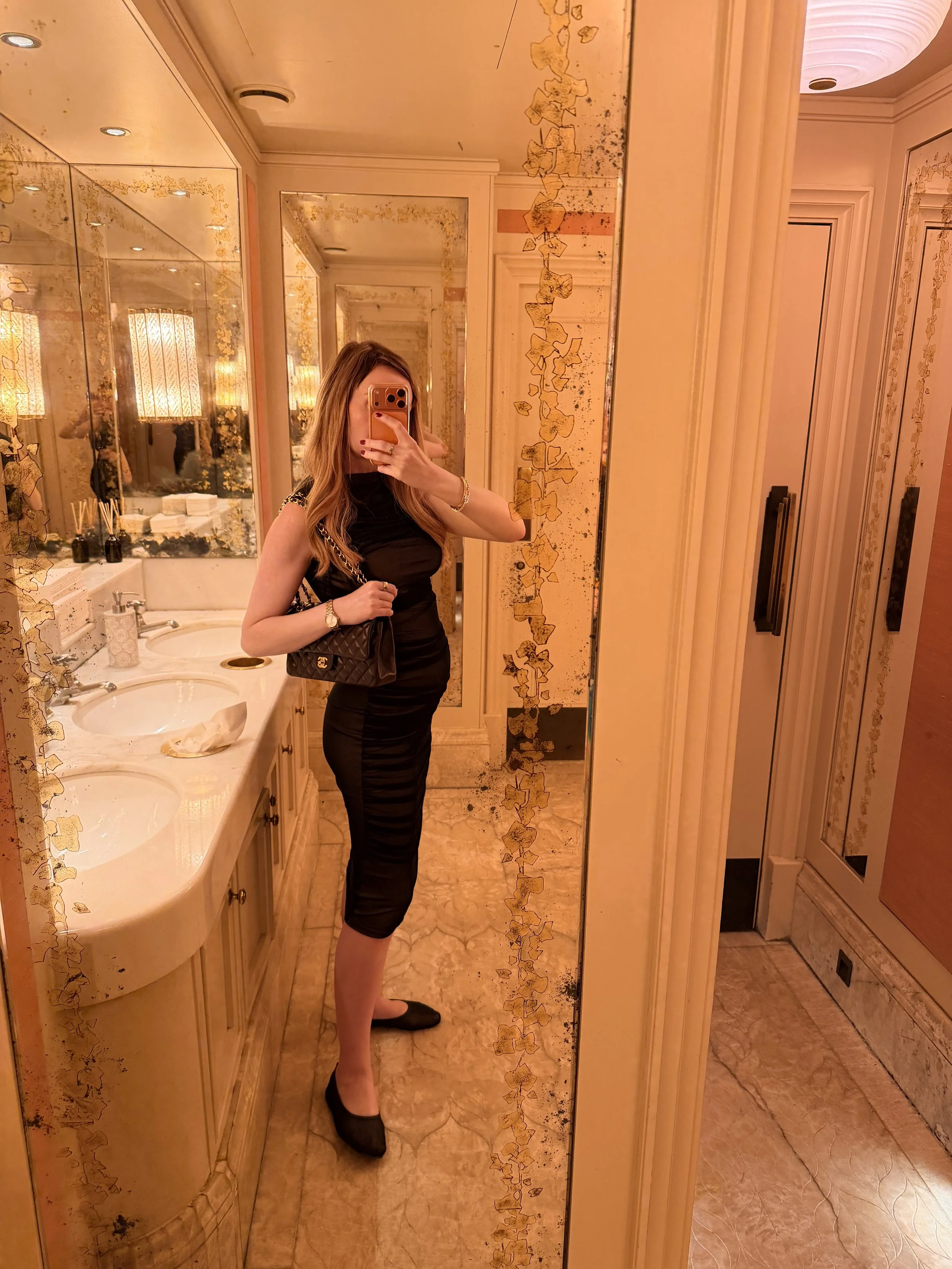 A pregnant independent escort in London taking a mirror selfie in a luxurious, gold-themed bathroom. She is wearing a black dress, black shoes, and carrying a small purse. The bathroom has elegant gold decorations and a marble vanity with two sinks.