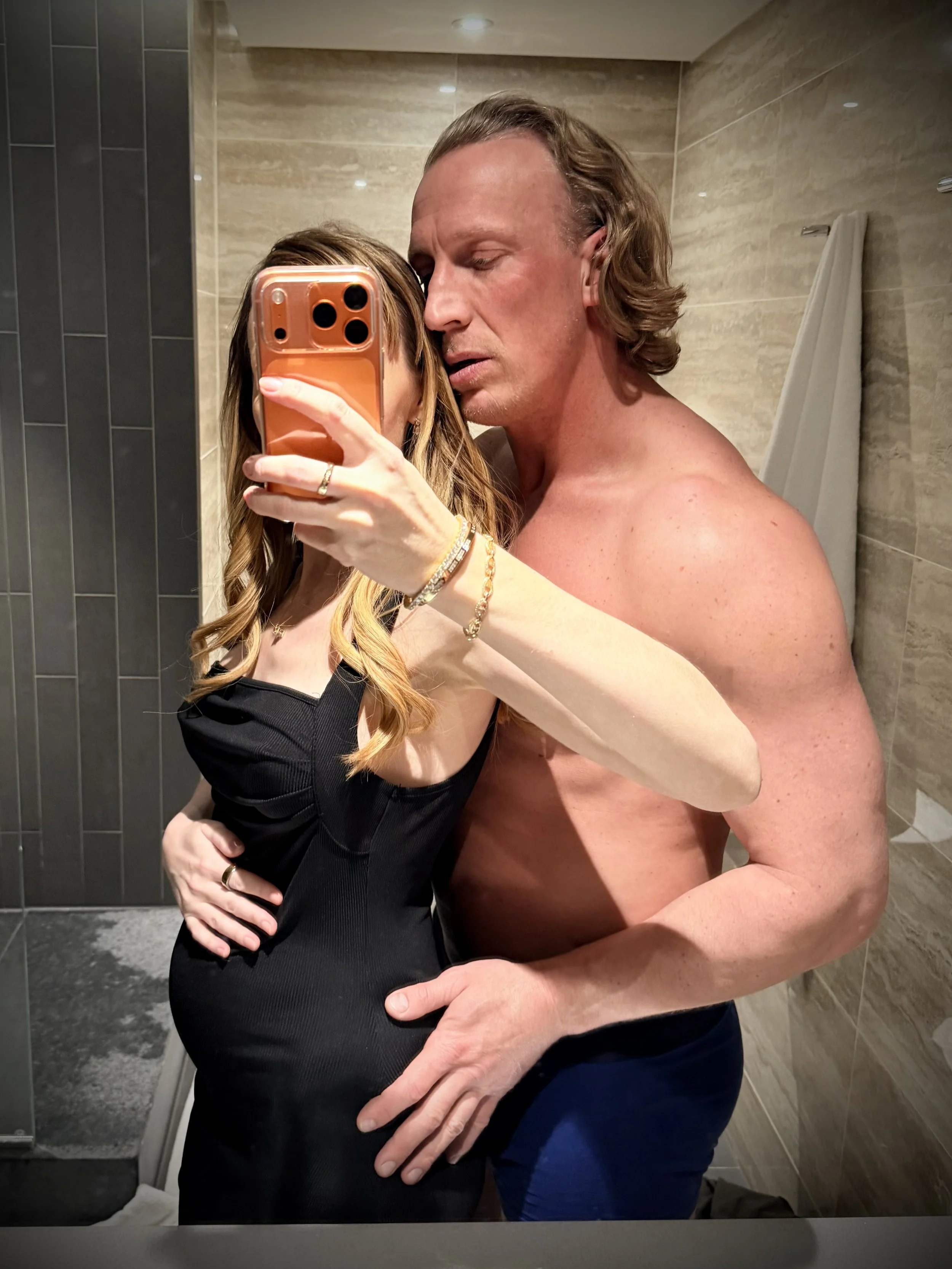 A pregnant escort and her cuckolding bull taking a mirror selfie in a London luxury hotel bathroom. The woman is wearing a black dress and holding the phone, while the bull is shirtless and standing close to her.