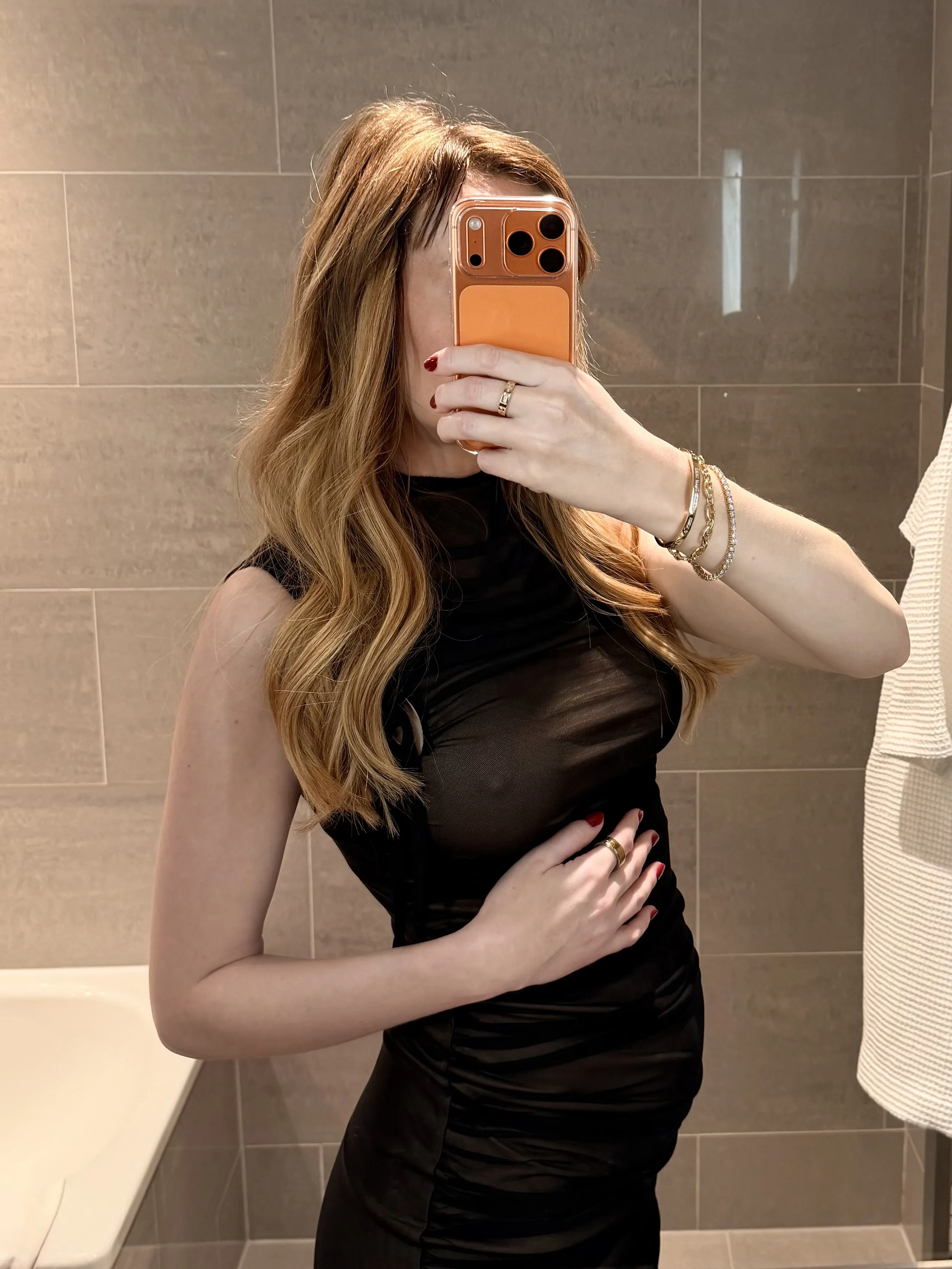 A luxury independent pregnant escort in London taking a mirror selfie in a bathroom, wearing a black dress, with long wavy hair, red nails, gold jewelry, and an orange phone case.