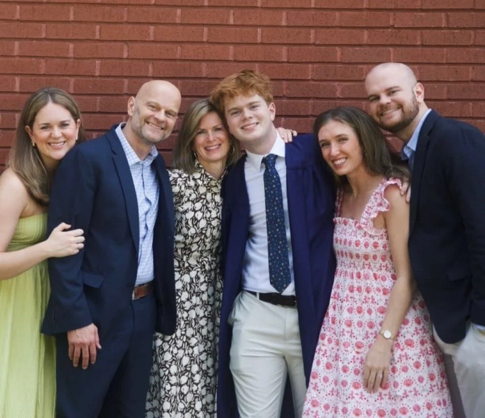 Group of six people celebrating, one person in a graduation gown, standing in front of a brick wall.