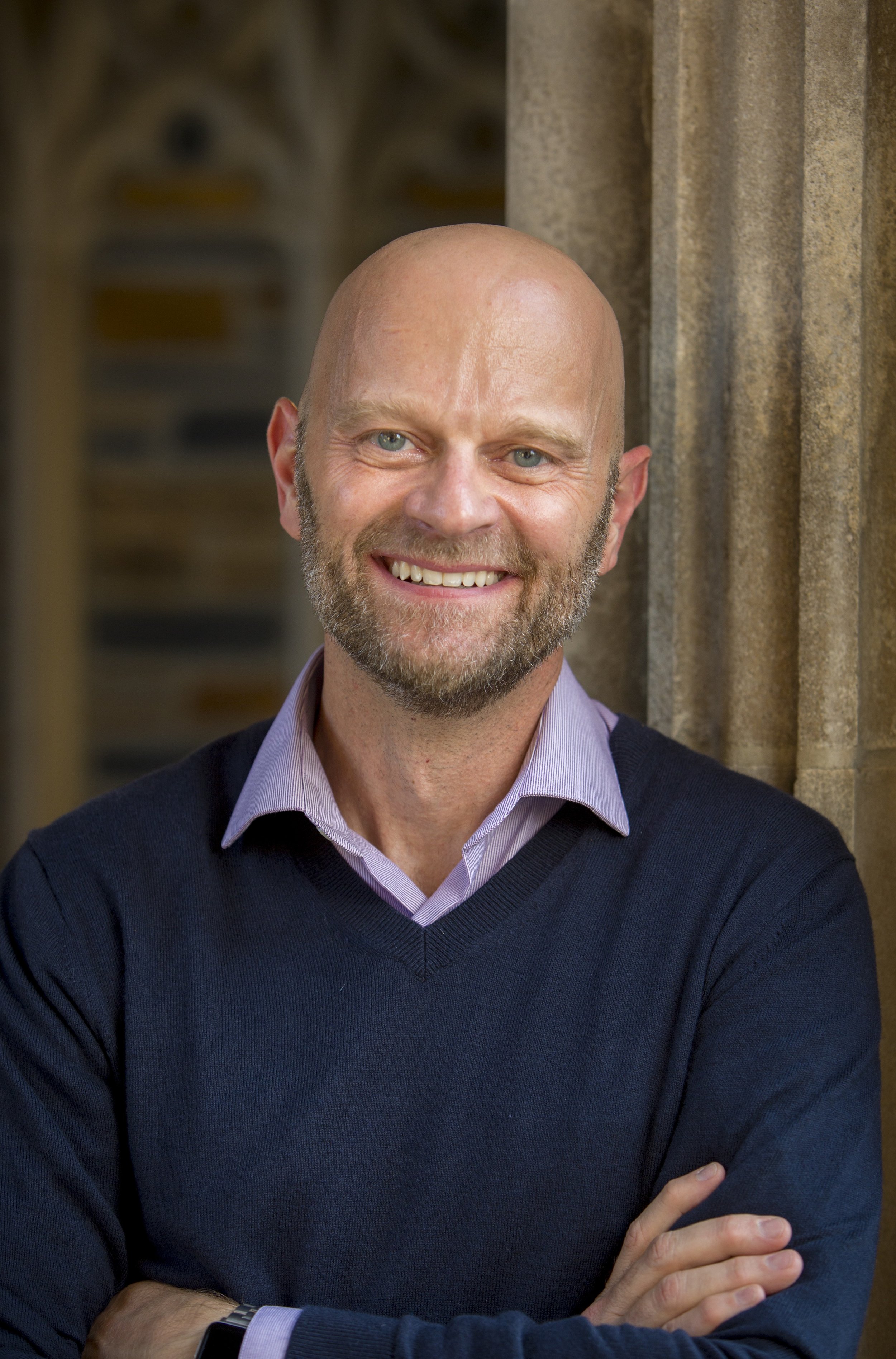 A smiling, bald man with a beard wearing a purple collared shirt and a dark blue sweater, standing outdoors against a stone wall, with arms crossed.
