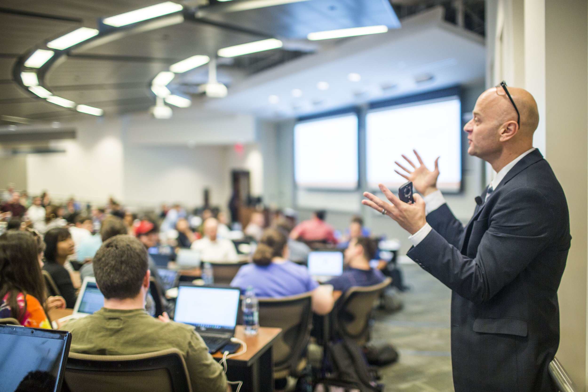 A man in a suit and glasses presenting to a conference room filled with attendees working on laptops.