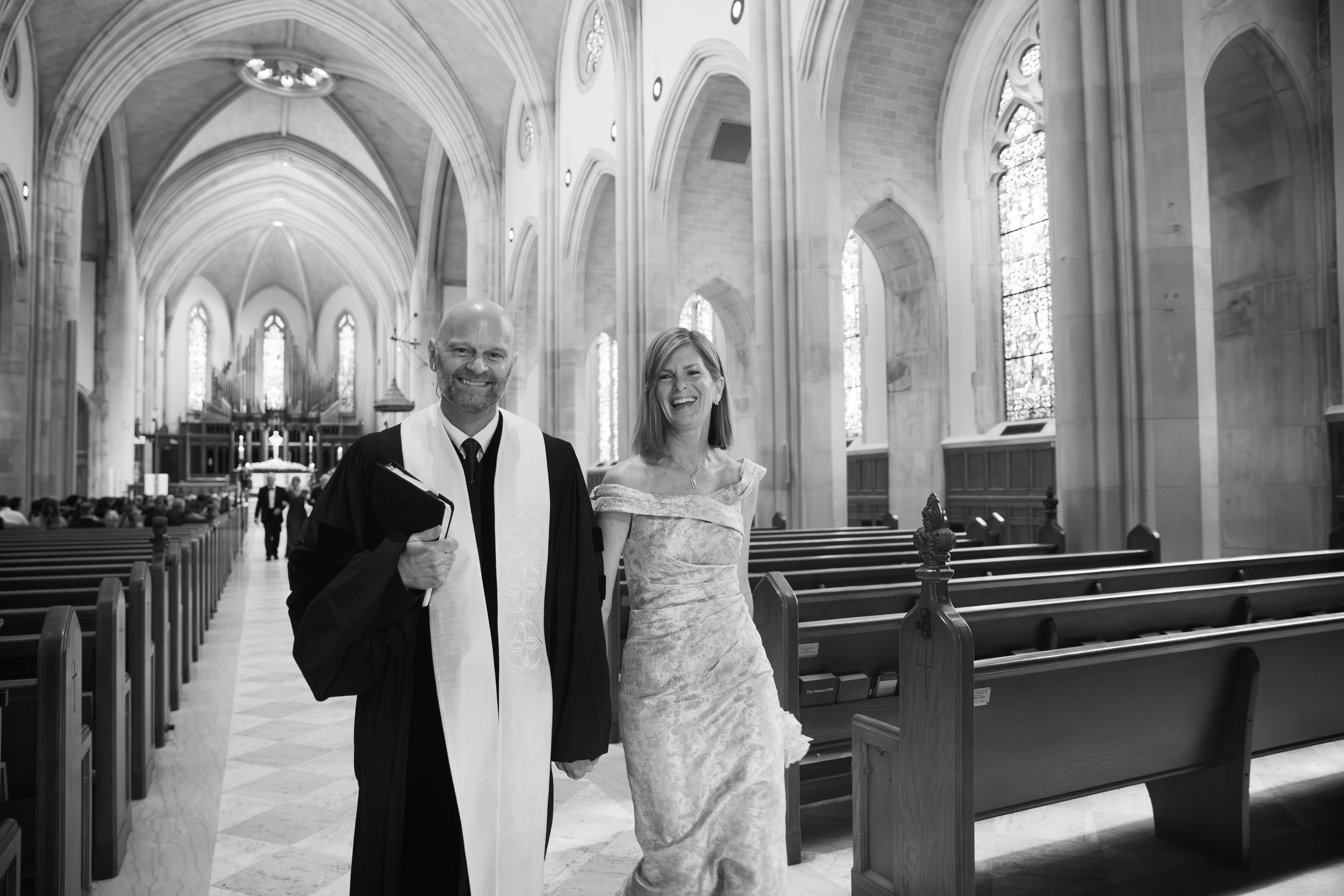A bride and groom standing inside a church, smiling and looking at the camera. The groom is wearing a black suit with a white clerical stole, holding a folder, and the bride is in a white gown with lace details, standing next to him. The church has high, arched ceilings, tall stained glass windows, and wooden pews.