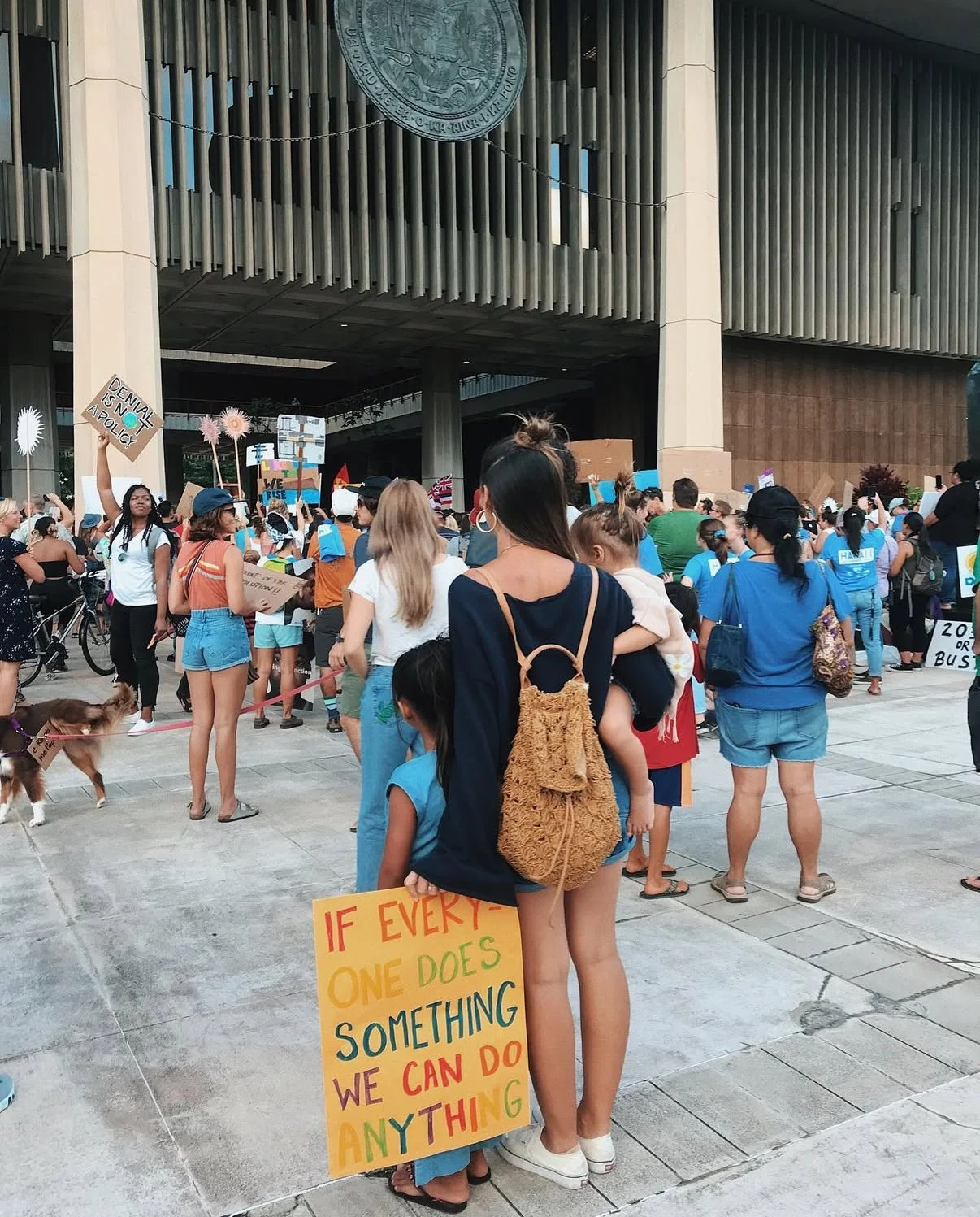 Crowd of people protesting outside a government building, holding signs and banners with messages about climate change and policy, with a woman holding a child and a colorful sign that reads, 'If every one does something we can do anything.'