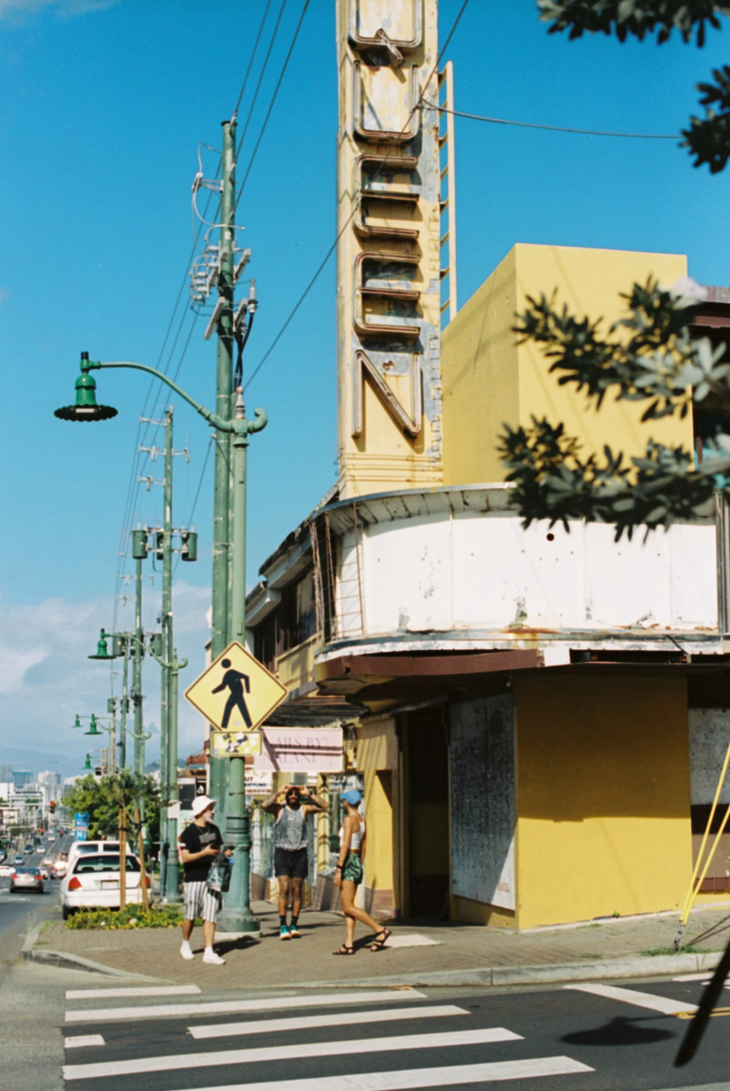 People standing at crosswalk in front of a weathered building with a vertical neon sign spelling 'GAMBLIN' and green street lamps, on a sunny day