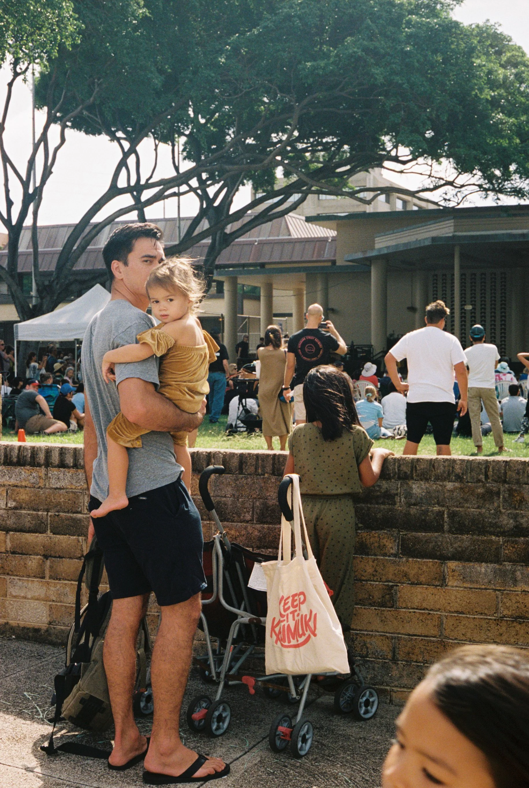 A man holding a young girl at a crowded outdoor event, with people sitting on the grass, a stage in the background, and trees overhead.