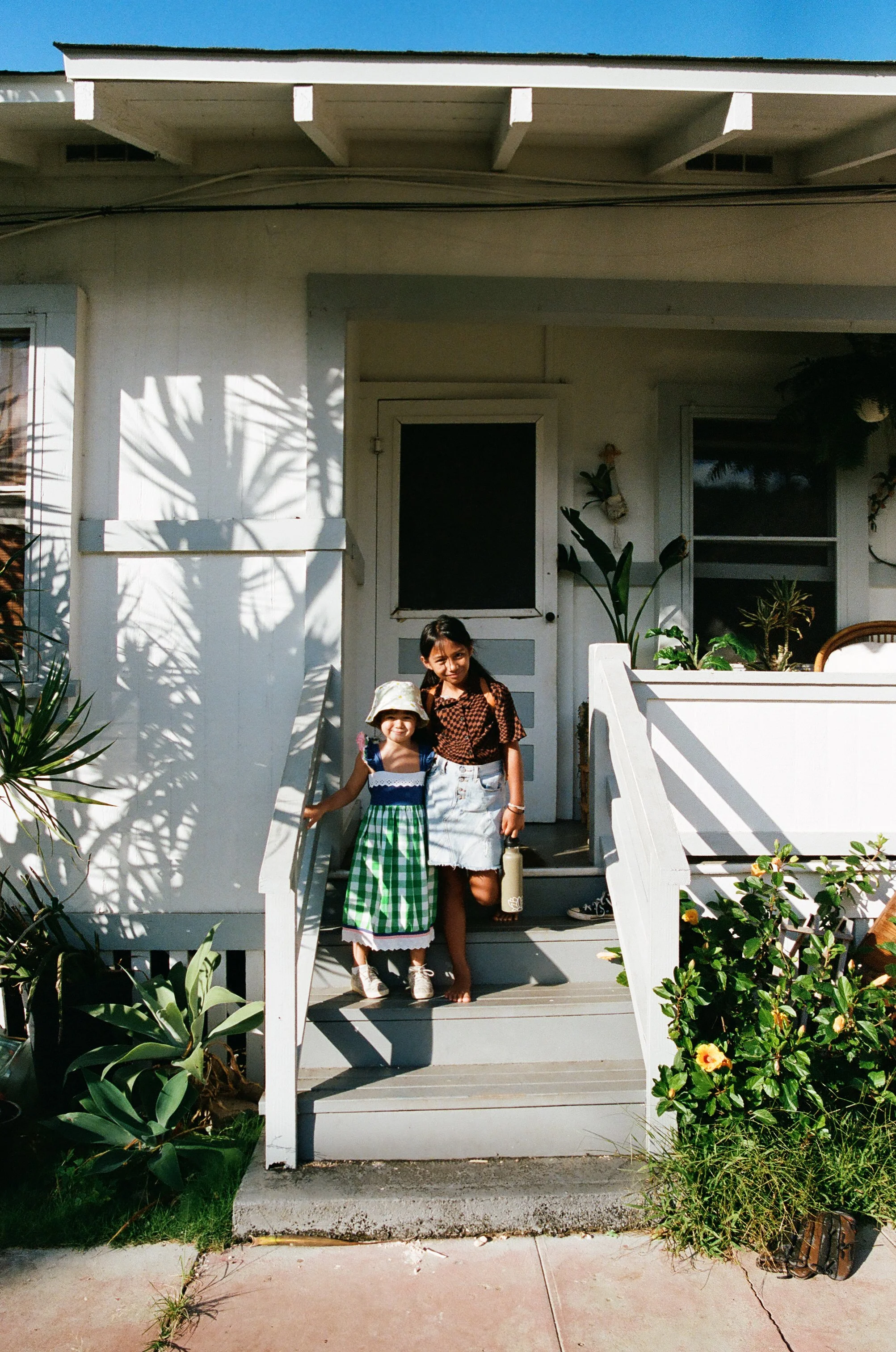 Two young girls standing on a porch of a white house with plants around, one girl wearing a green checkered dress and a sun hat, the other girl wearing a brown patterned shirt and a denim skirt.