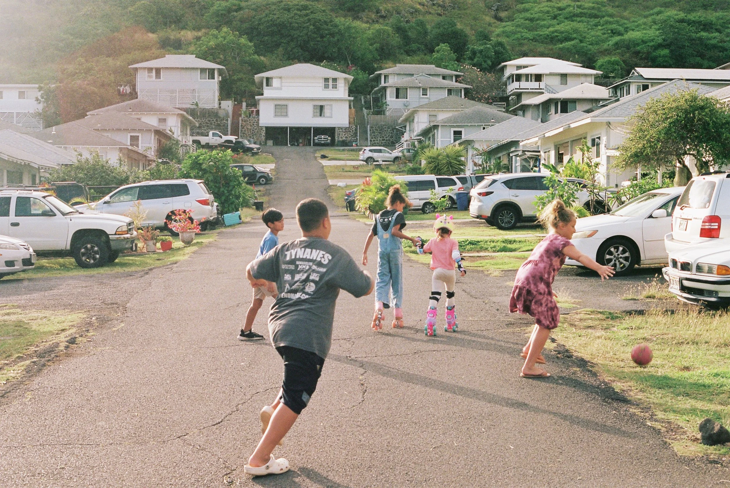 Children playing outdoor in a residential neighborhood, some on rollerblades, a girl kicking a ball, surrounded by parked cars and houses on a hilly street with greenery.