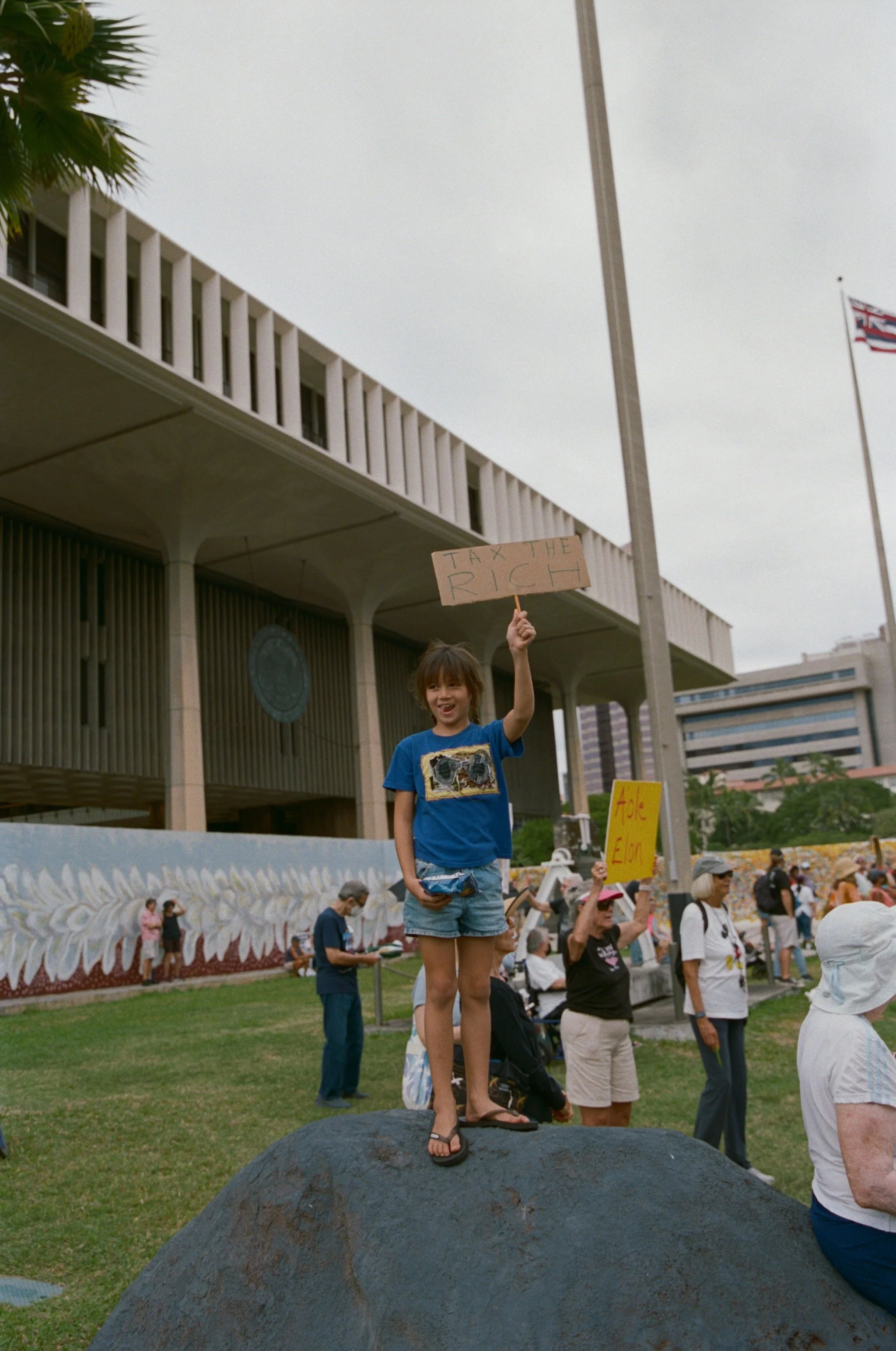 Young girl standing on a rock, holding a sign that says "Tax the Rich," during a protest or rally in an urban area.