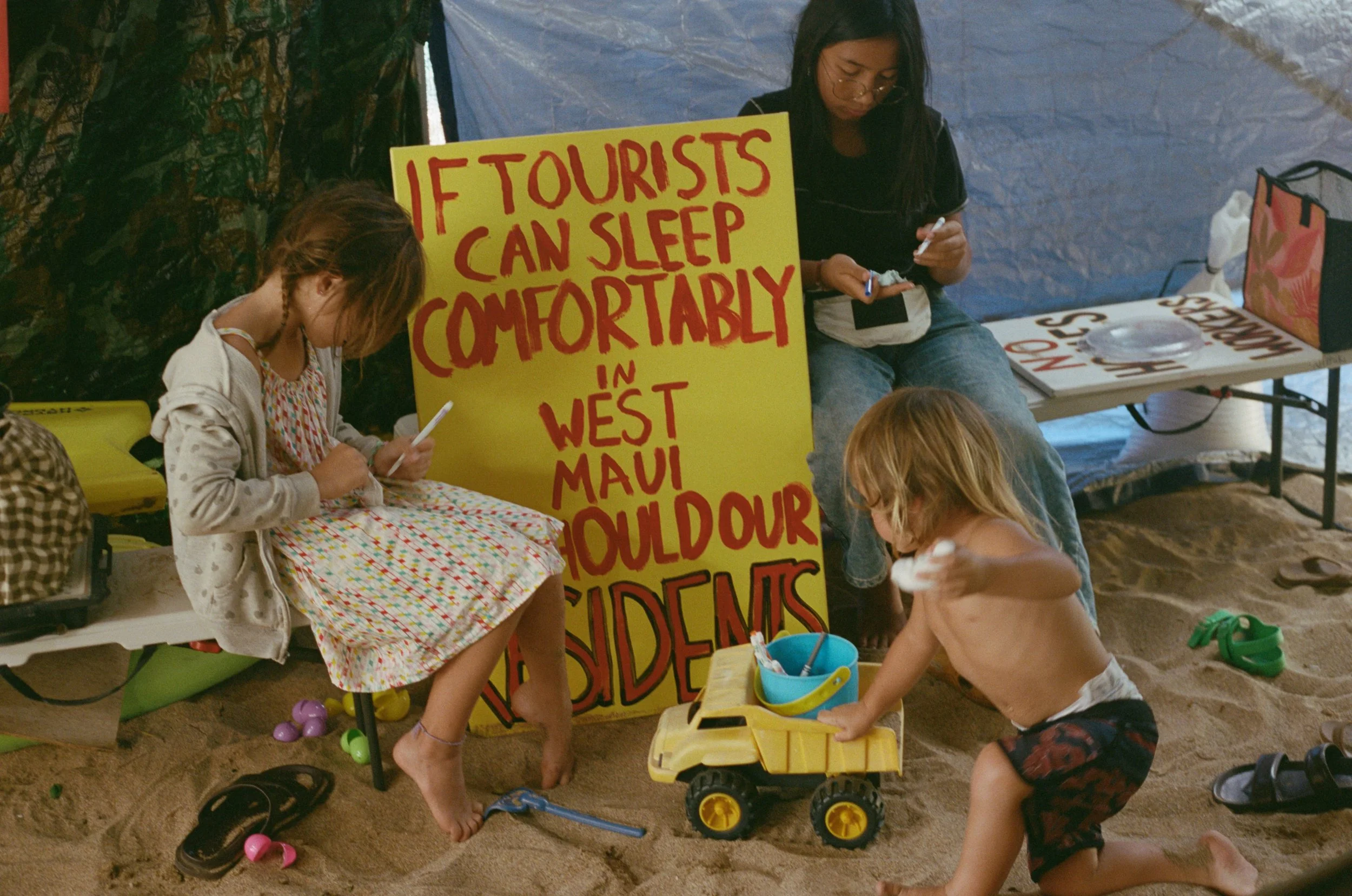 Children playing and writing on a sandy beach, with a large yellow sign that reads, "If tourists can sleep comfortably in West Maui, Oahu, and Dour residents".