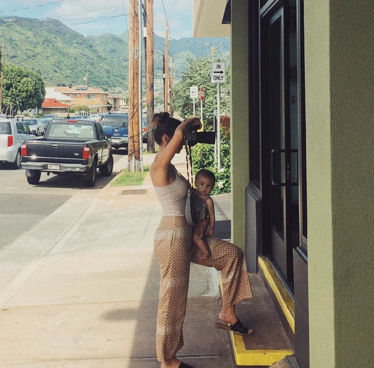 Woman taking a photo with a camera, with a child sitting on her leg, standing outside a building on a sidewalk with parked cars and a mountain behind in the background.