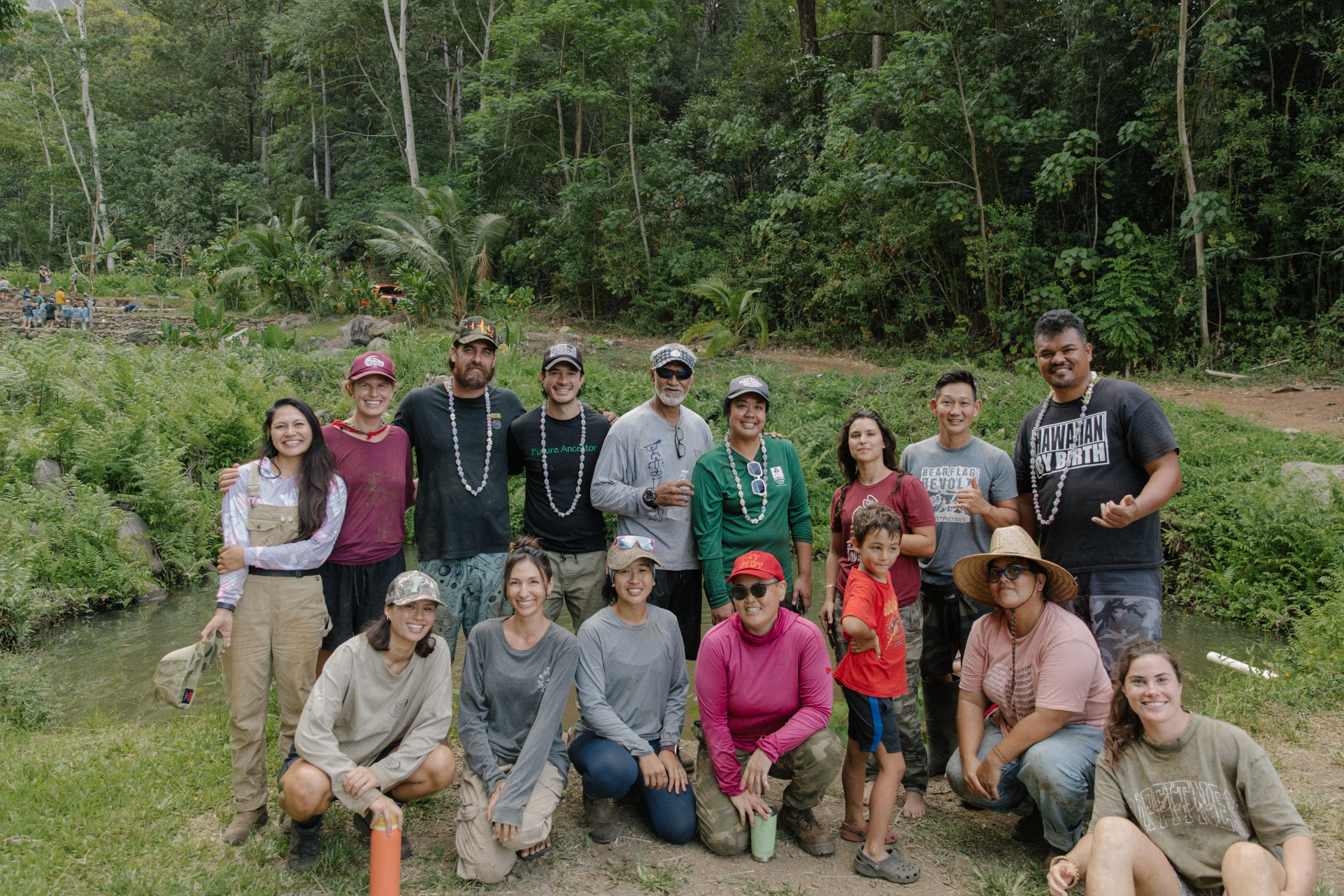 Group of people outdoors in a lush green area, some wearing leis, posing for a photo with trees in the background.