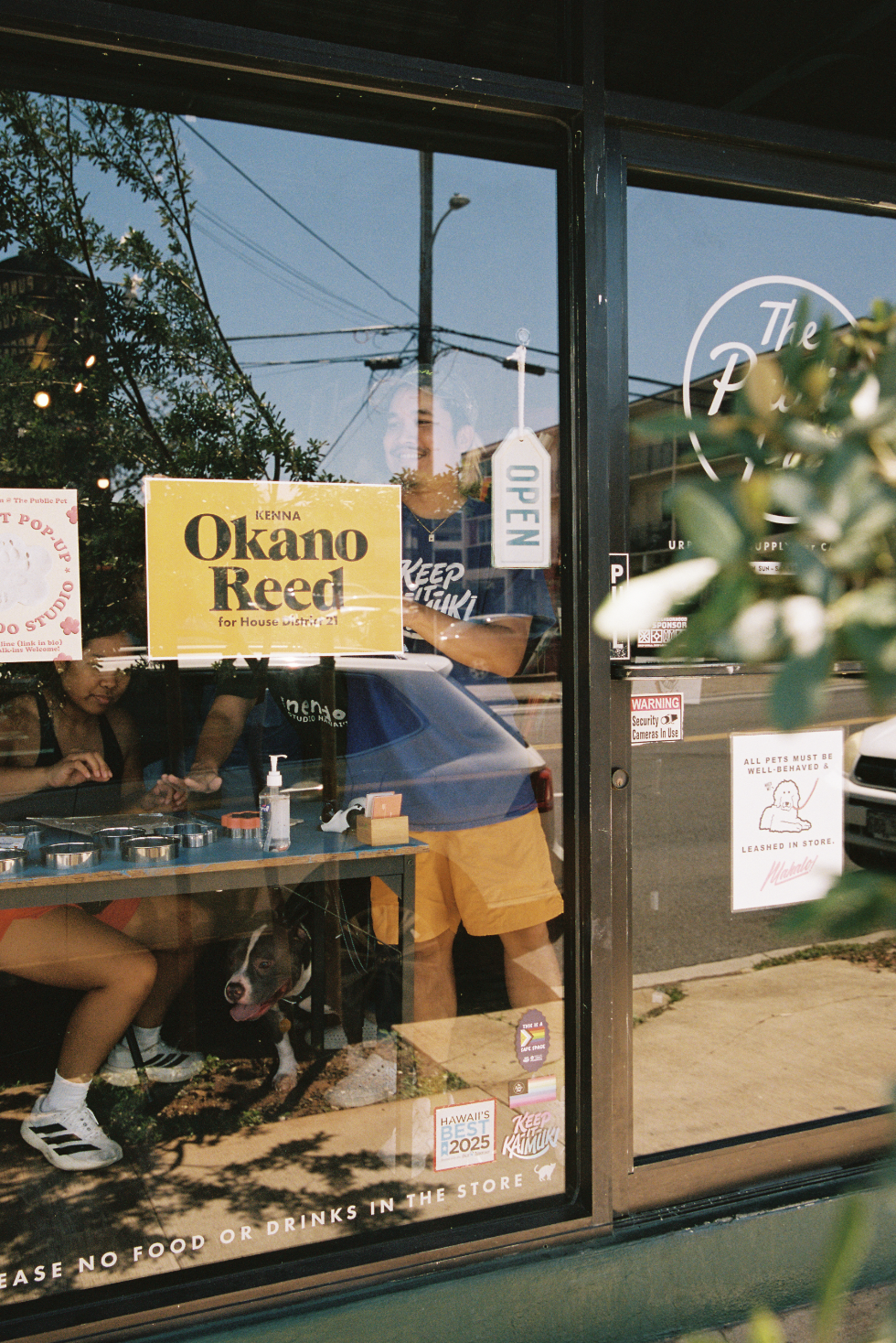 View through the window of a pet store or café showing a person smiling inside, with a dog sitting on the floor. There are signs on the window, including one that says 'Kenna Okano Reed for House District 21' and another with an 'Open' sign hanging. Outside, cars and a street with overhead power lines are visible.