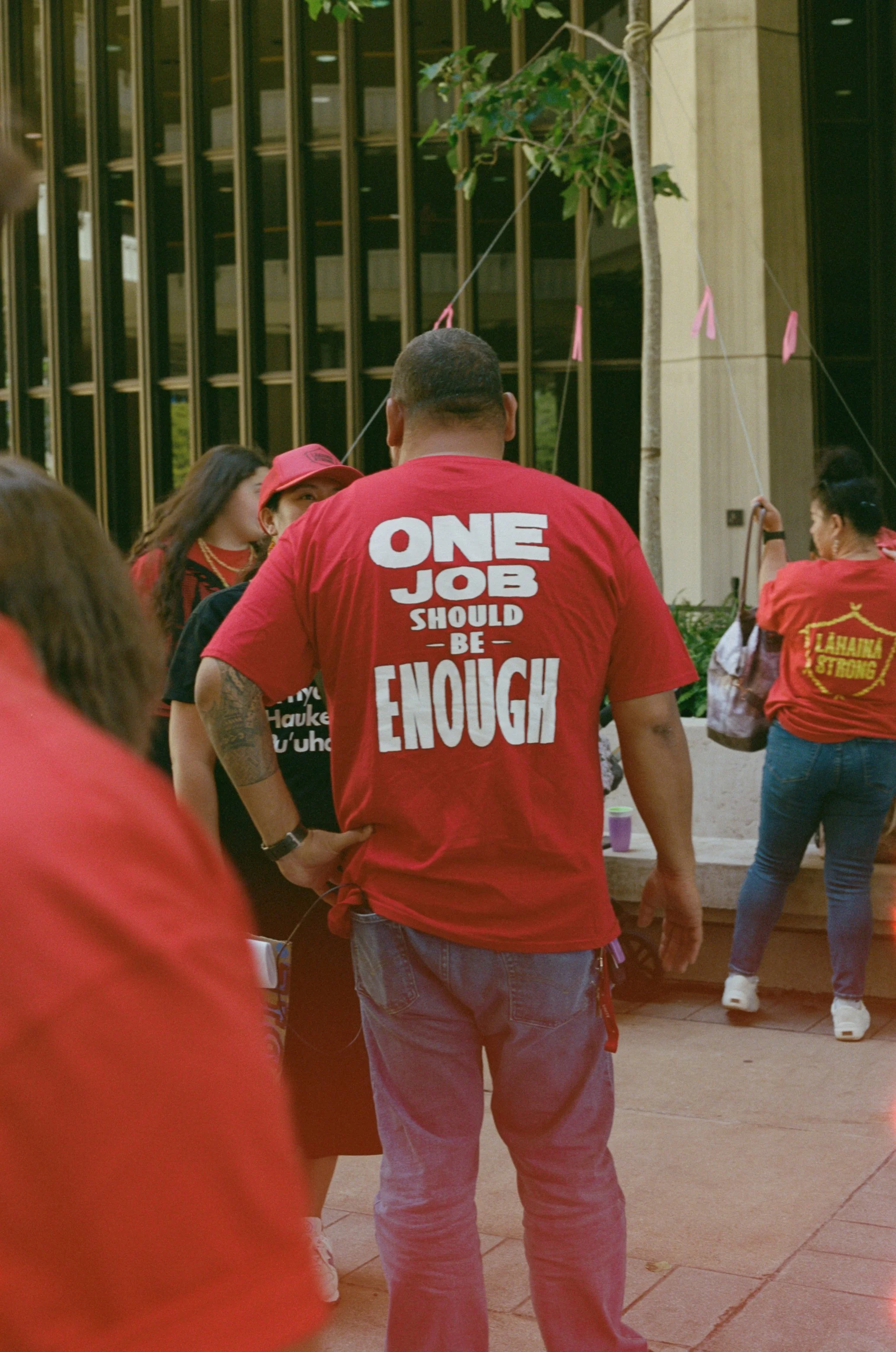 A man wearing a red T-shirt with the text 'One Job Should Be Enough' stands in a group of people, some dressed in red, outdoors near a building with glass windows and a tree.