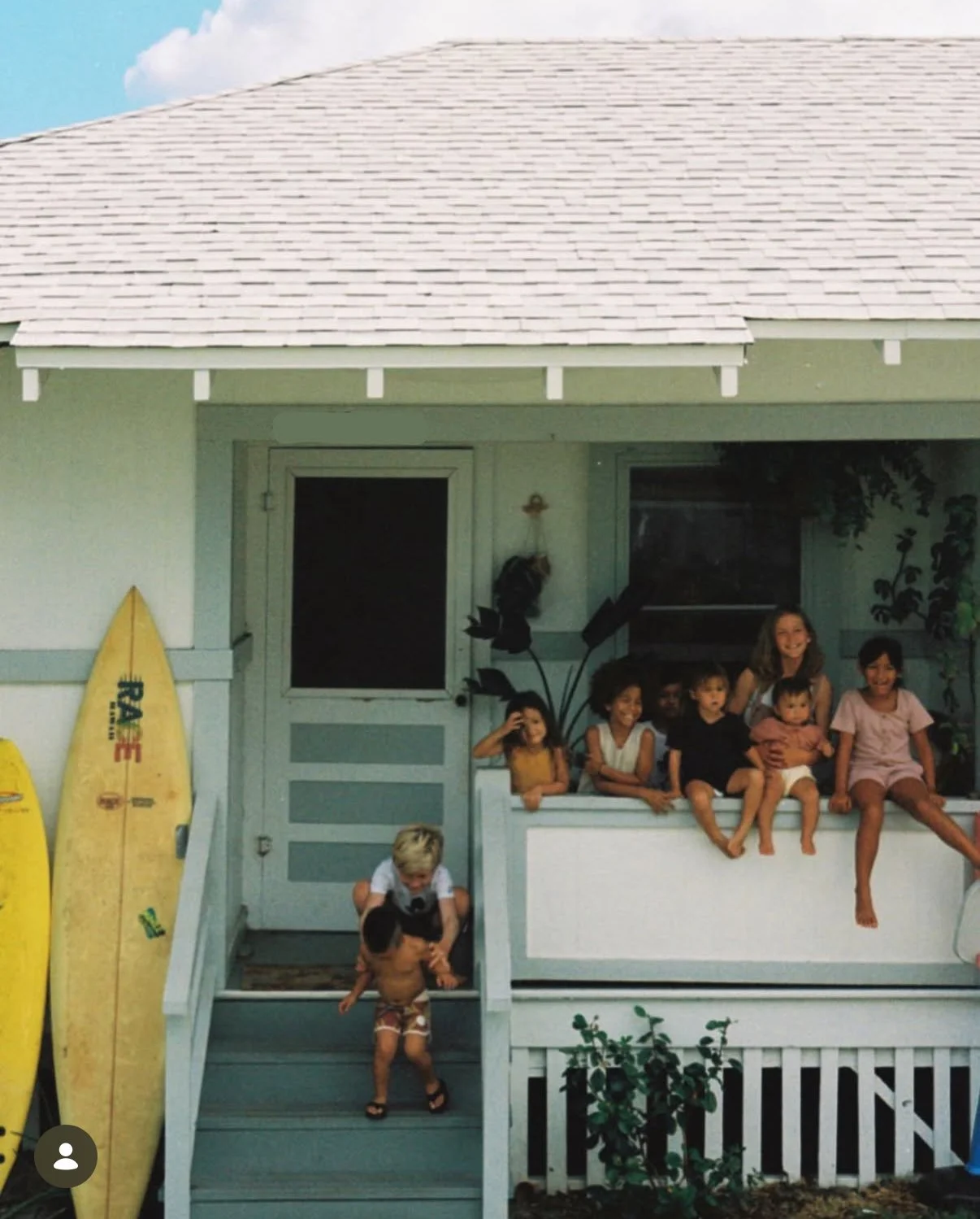 Children sitting on the porch of a house with surfboards leaning against the wall, some children playing on the stairs, and others sitting on the porch, smiling.