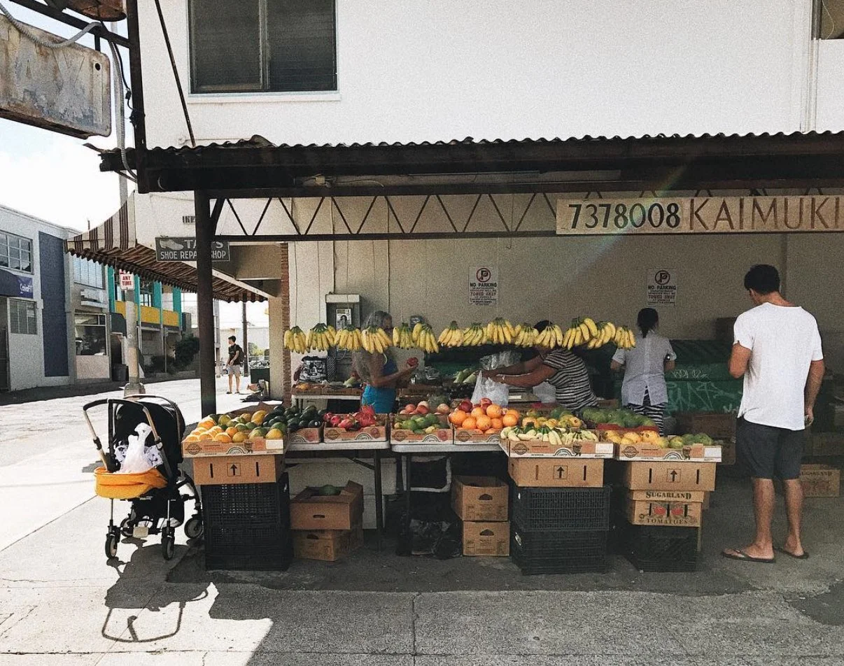 Street vendor fruit stand with bananas, oranges, and melons, and a stroller nearby, in an outdoor shopping area.