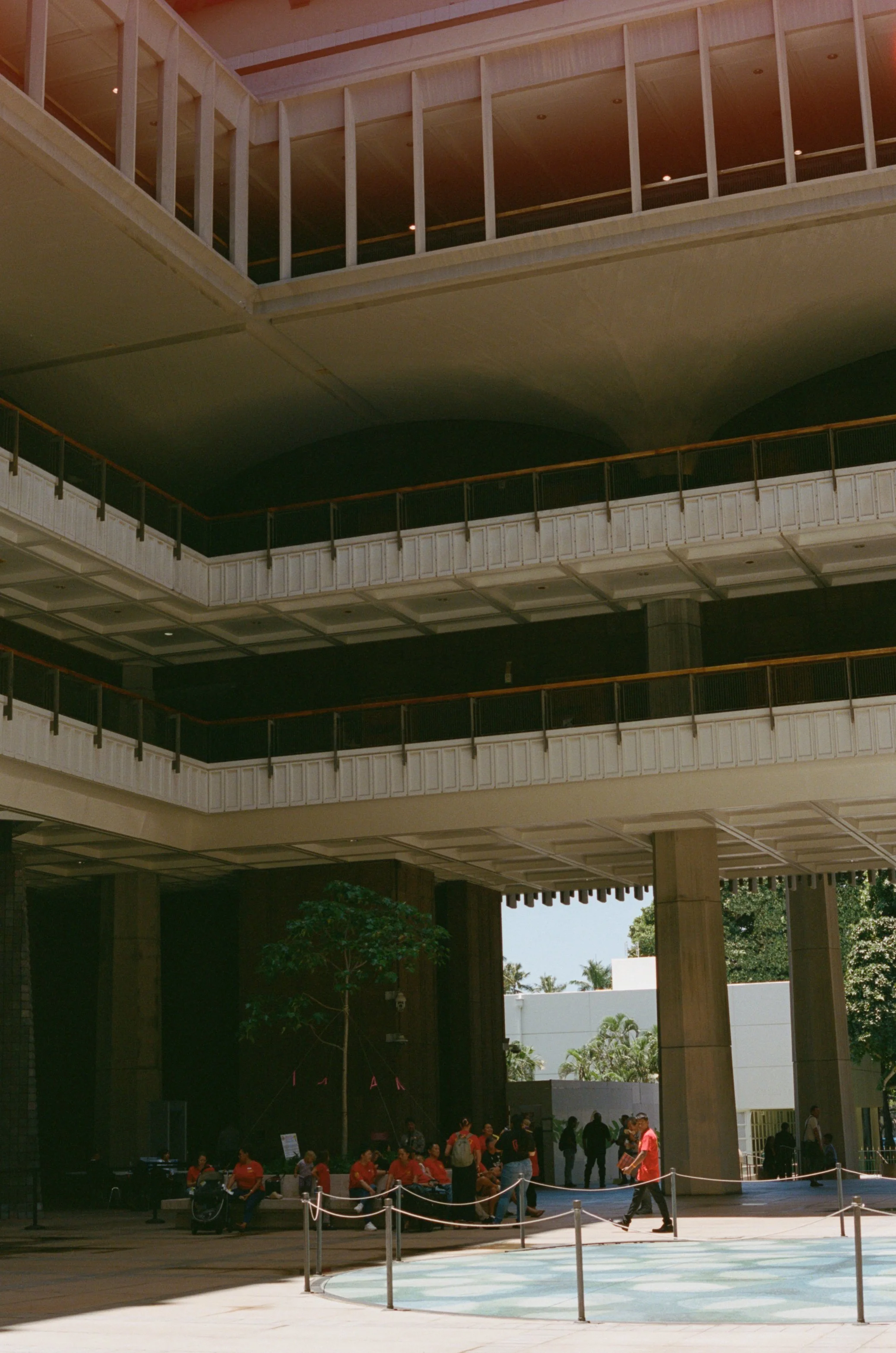 People in red shirts sitting and standing in front of a large building with multiple floors, columns, and trees, possibly at an event or gathering.
