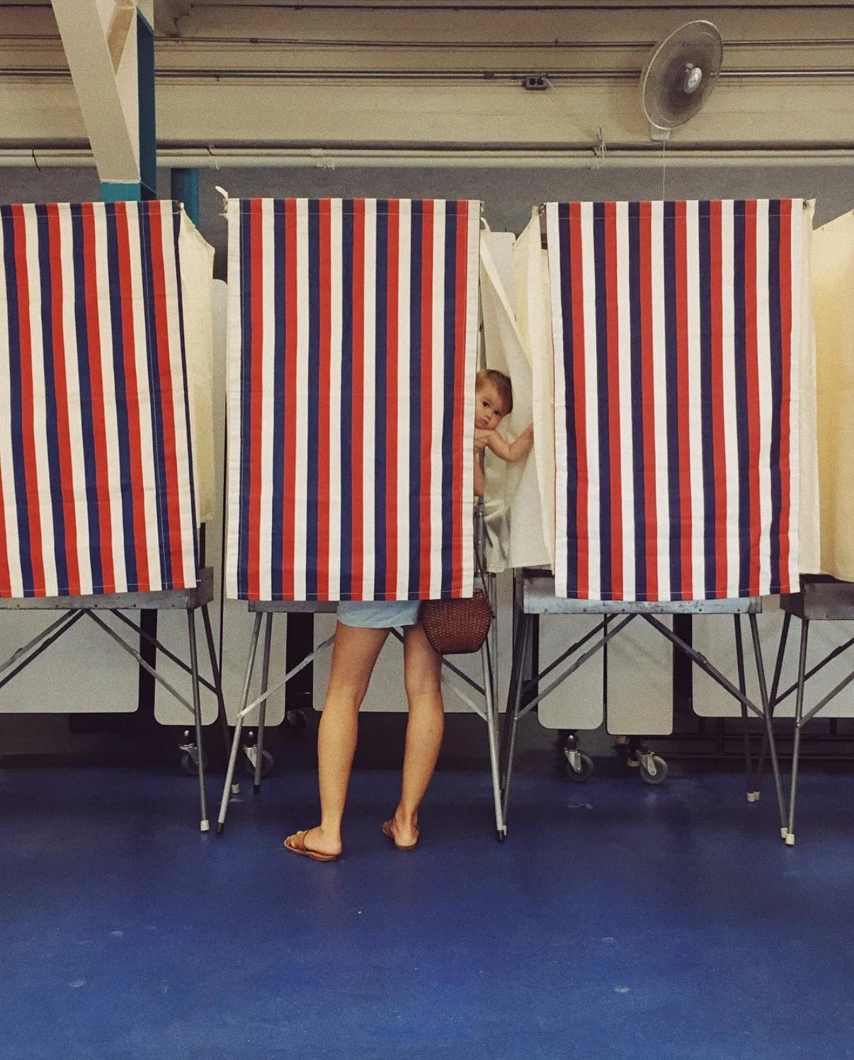 Child peeking out from a striped voting booth with a person standing behind, only their legs and feet visible.