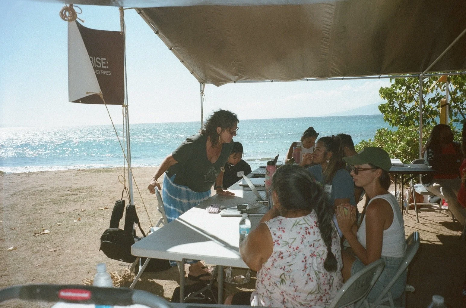 Group of people attending outdoor meeting under a tent on a beach, with laptops, notebooks, and water bottles, near the ocean with a view of the horizon.