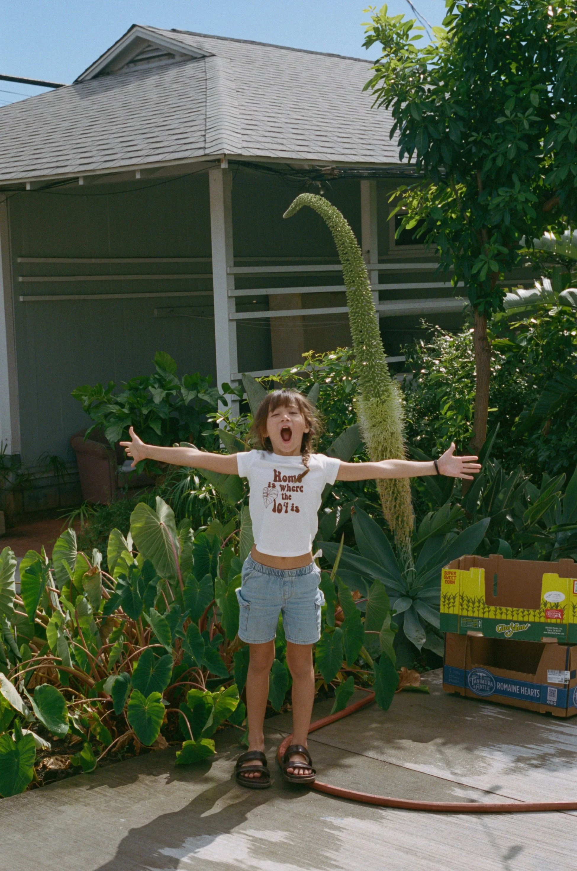 Young girl with arms outstretched, standing on a sidewalk in front of lush green plants, in a garden area, with a house background and a large flowering plant behind her.