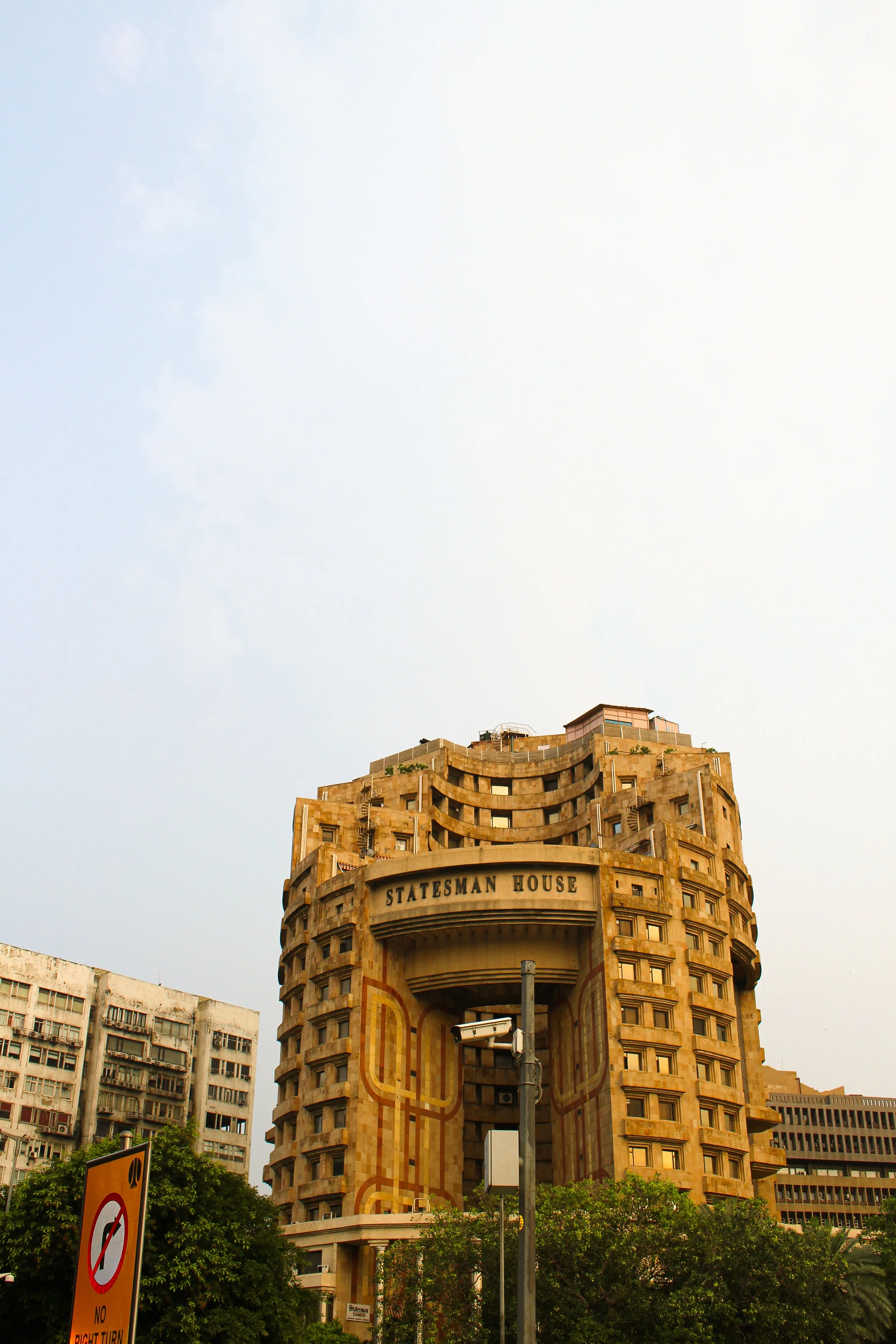 The image shows the Estateman House building, a tall, brown, art deco style structure with curved design elements, situated in an urban area with neighboring buildings, street signs, and greenery.