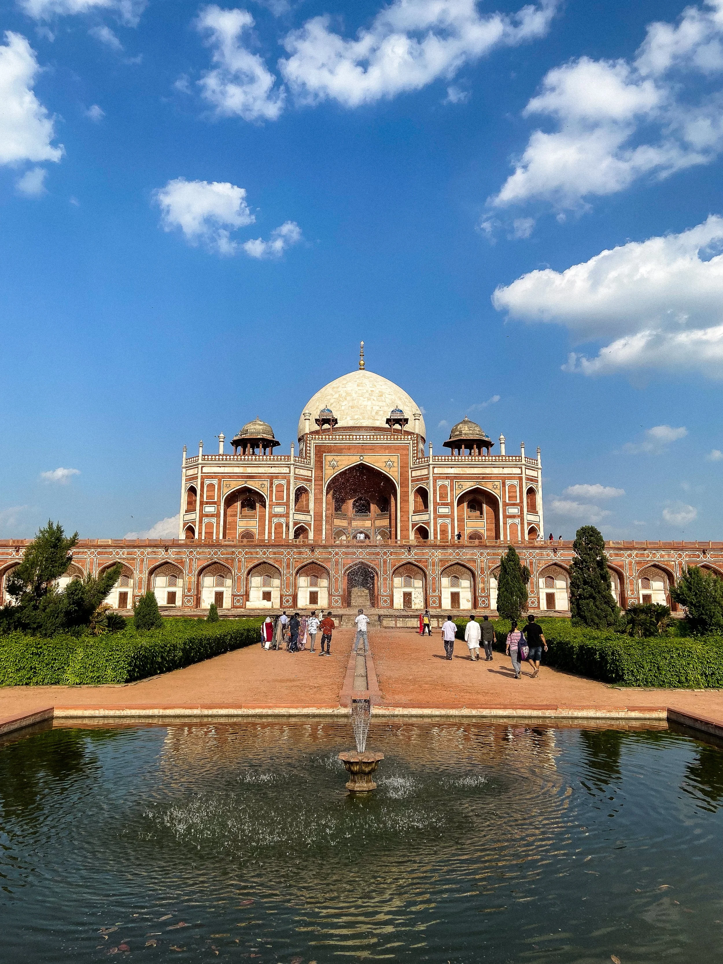 The Taj Mahal, an iconic white marble mausoleum with a large central dome and surrounding smaller domes, is viewed from the front with a water fountain in the foreground. Visitors are walking along the pathway leading to the entrance, with a clear bl