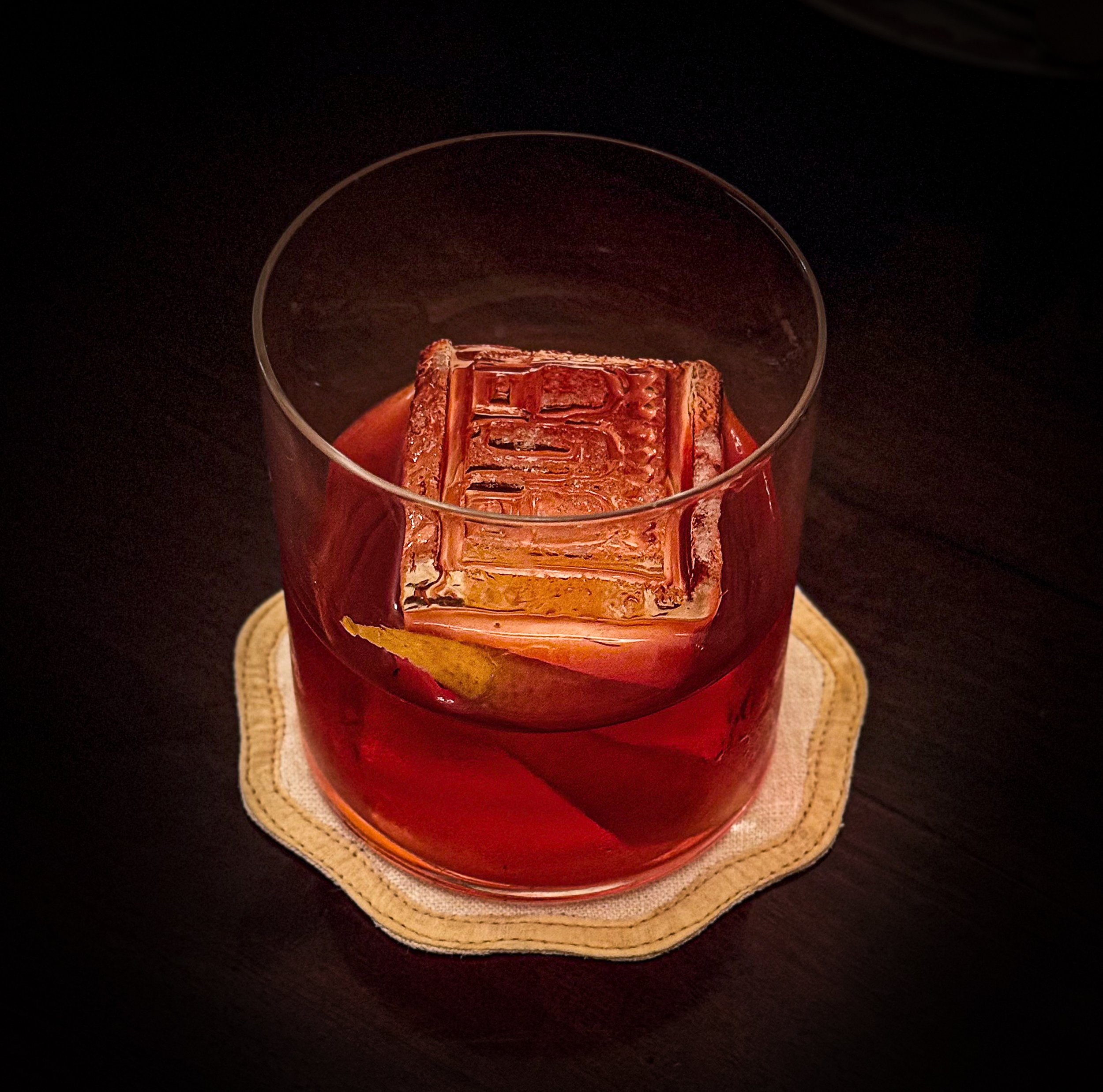 A glass of red cocktail with a large ice cube and a lemon wedge on a cloth coaster on a dark surface.