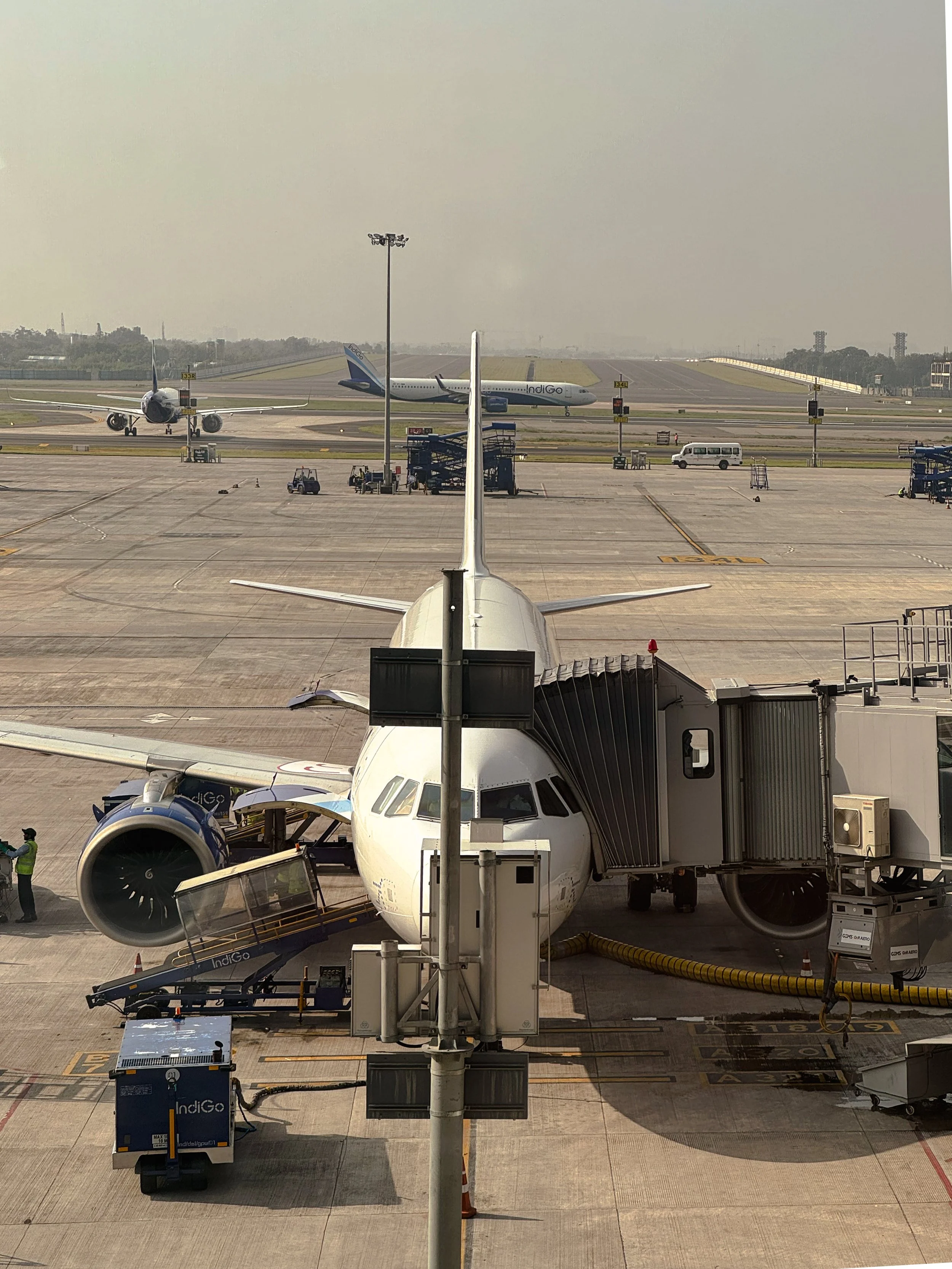 View of an airport terminal with a white airplane parked at the gate, connected to the jet bridge. In the background, two airplanes are on the runway, one with an IndiGo logo. Ground crew and luggage carts are visible around the aircraft, with a hazy