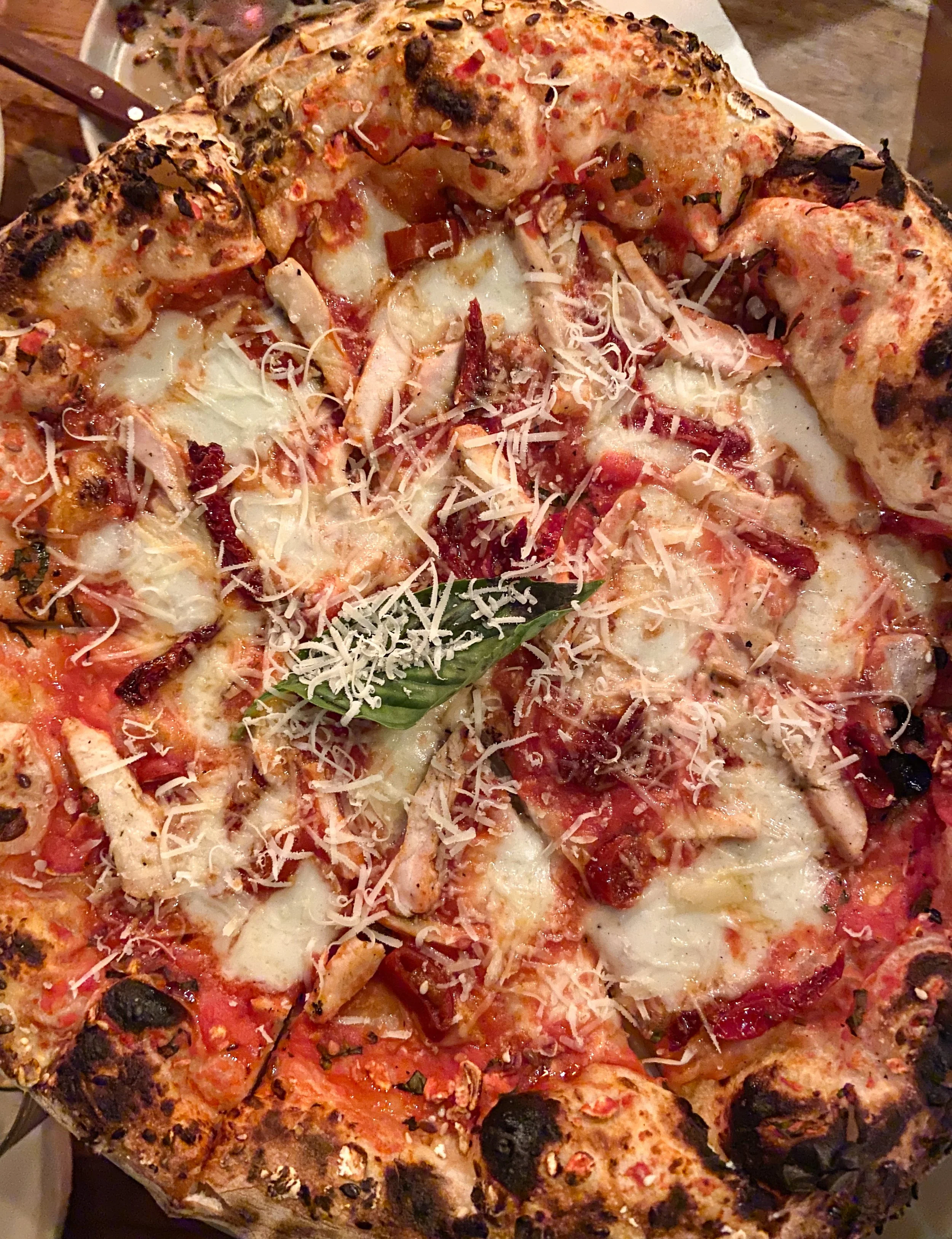 Close-up of a pizza with cheese, tomato sauce, chicken, bacon, basil, and various toppings on a wood table.