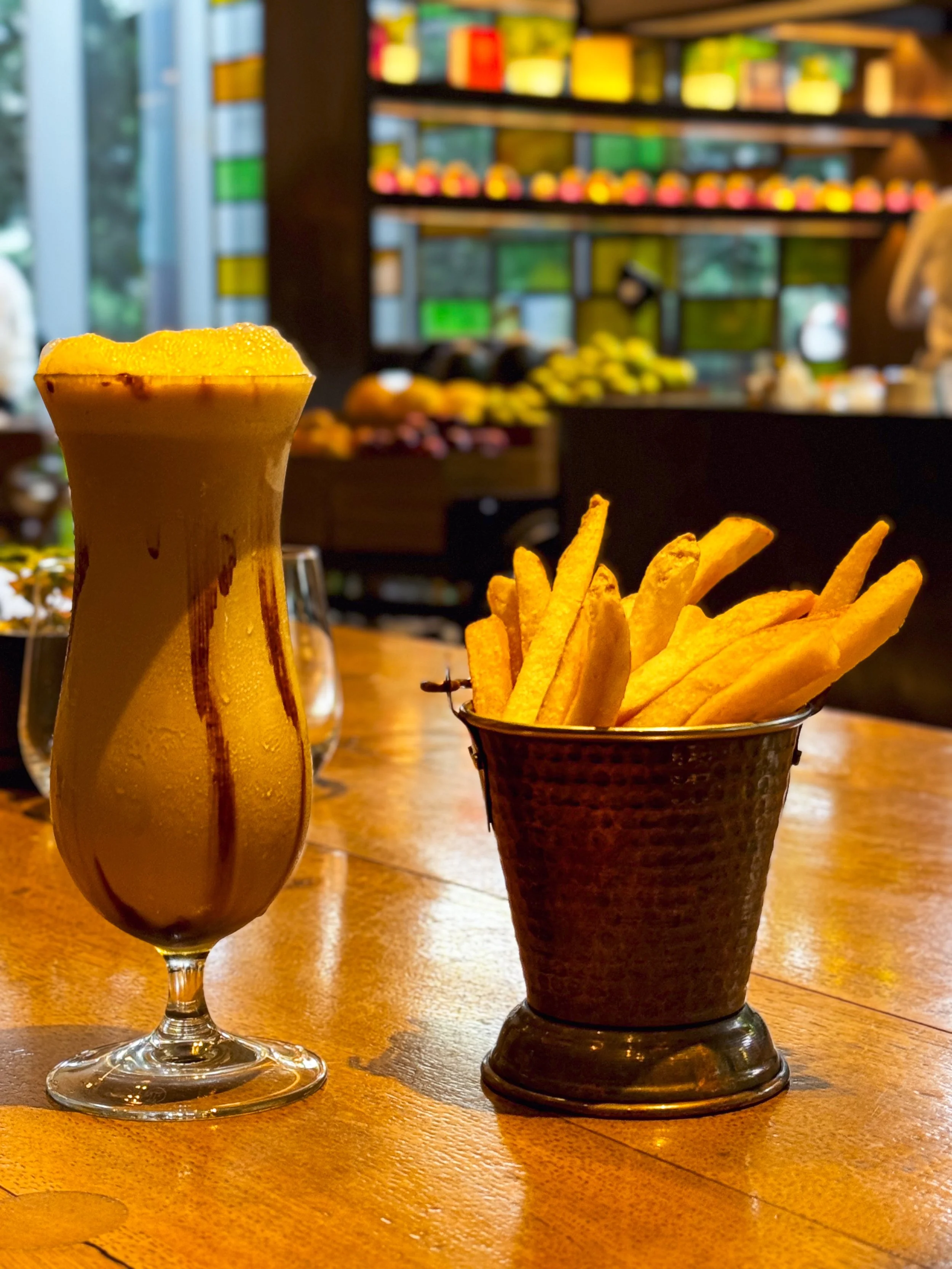 A glass of a peach-colored cocktail with a foamy top and a drizzle of chocolate syrup, alongside a metal cup filled with French fries, on a wooden table inside a restaurant or café with a colorful blurred background.