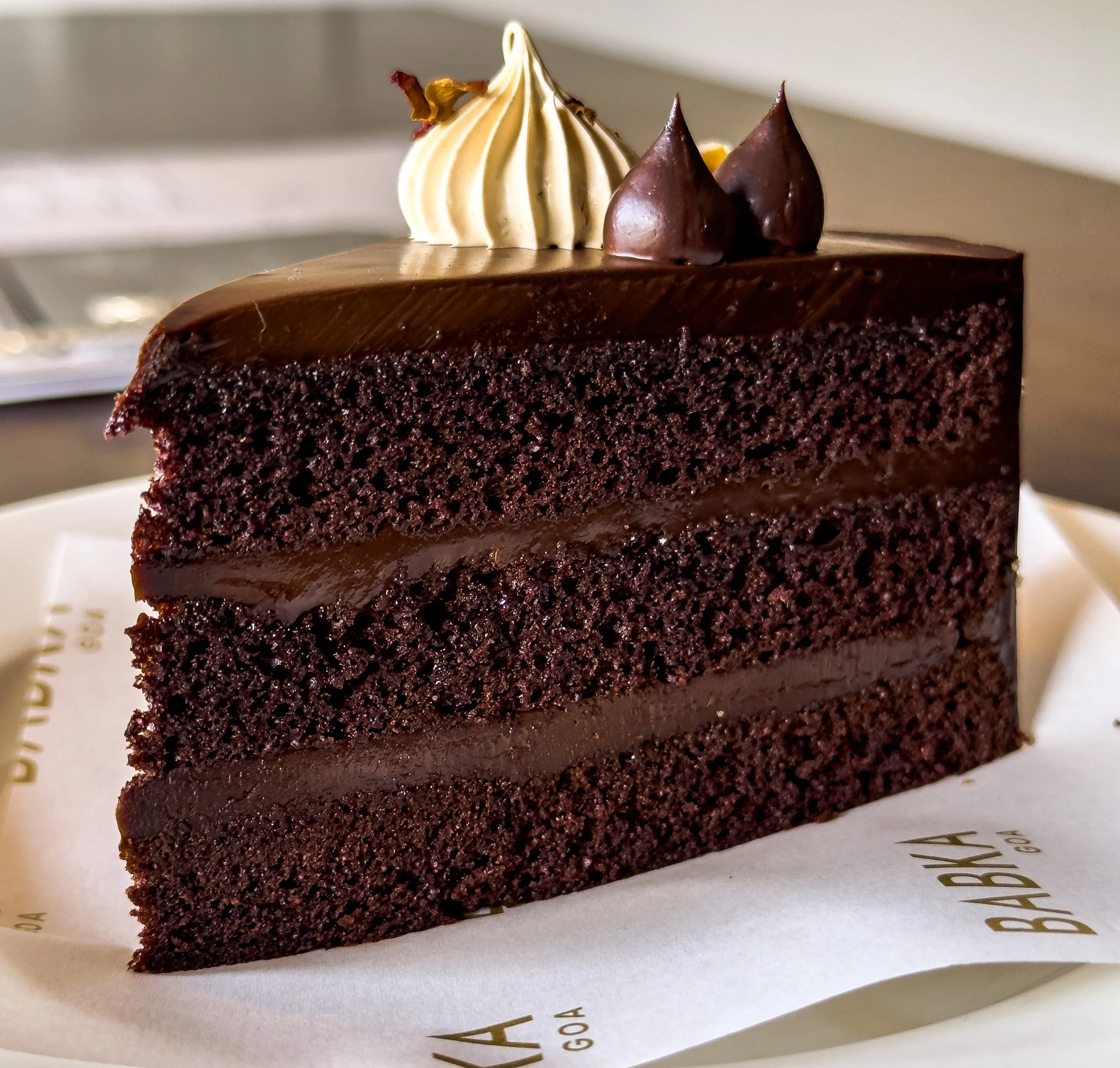 A slice of rich chocolate cake with three layers, topped with chocolate and white cream dollops.