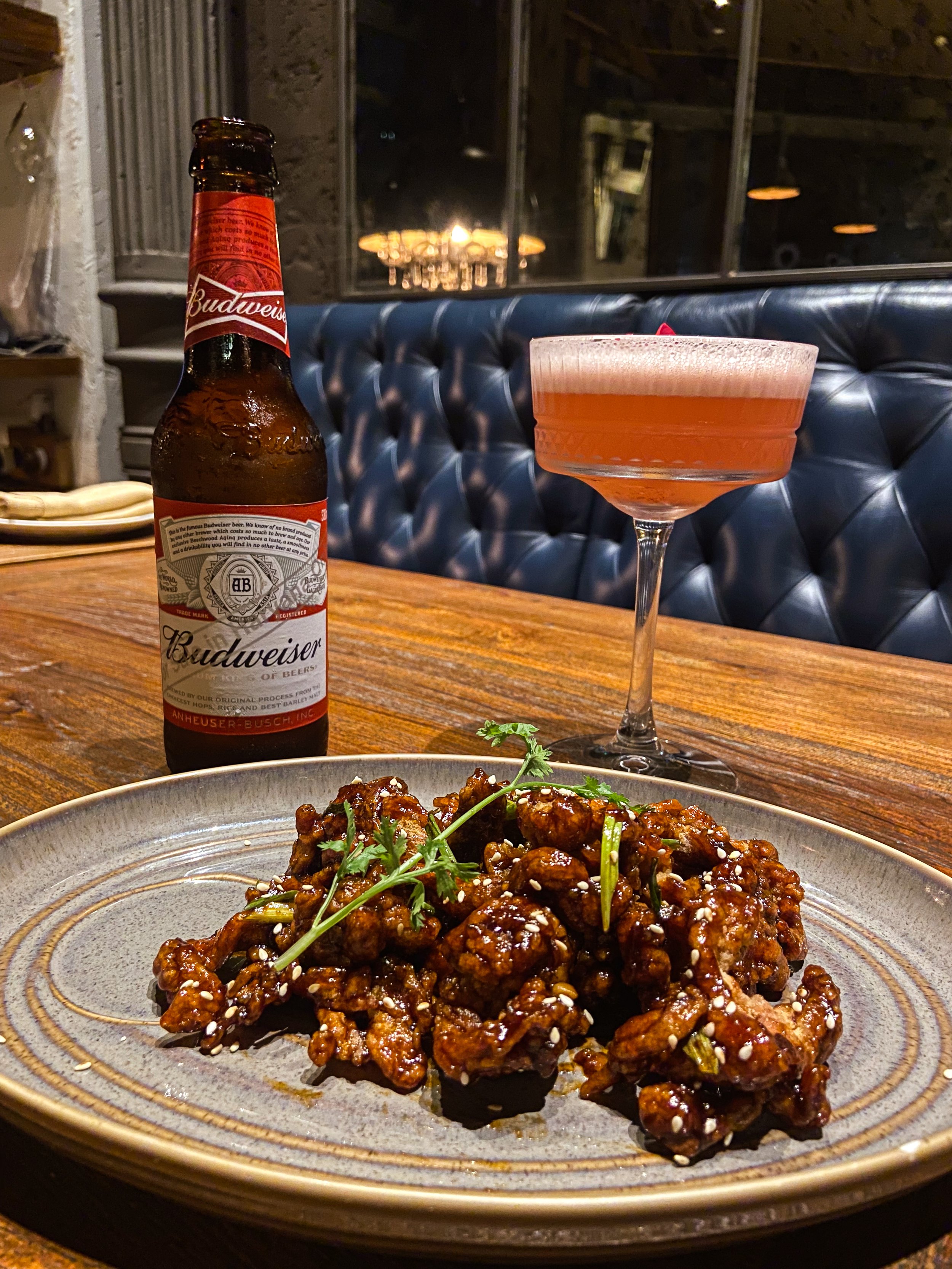 Plate of Asian-style glazed chicken garnished with cilantro, a Budweiser beer bottle, and a cocktail glass containing a pinkish drink on a wooden table in a cozy restaurant setting.